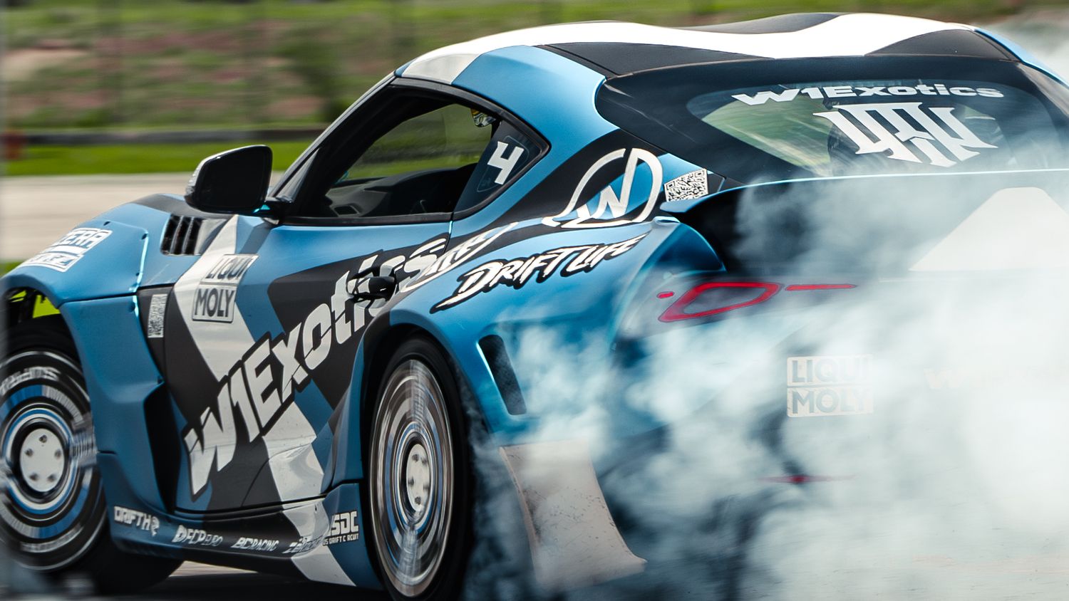 A blue drift car with W1 Exotics livery performs a smoky burnout on a race track.