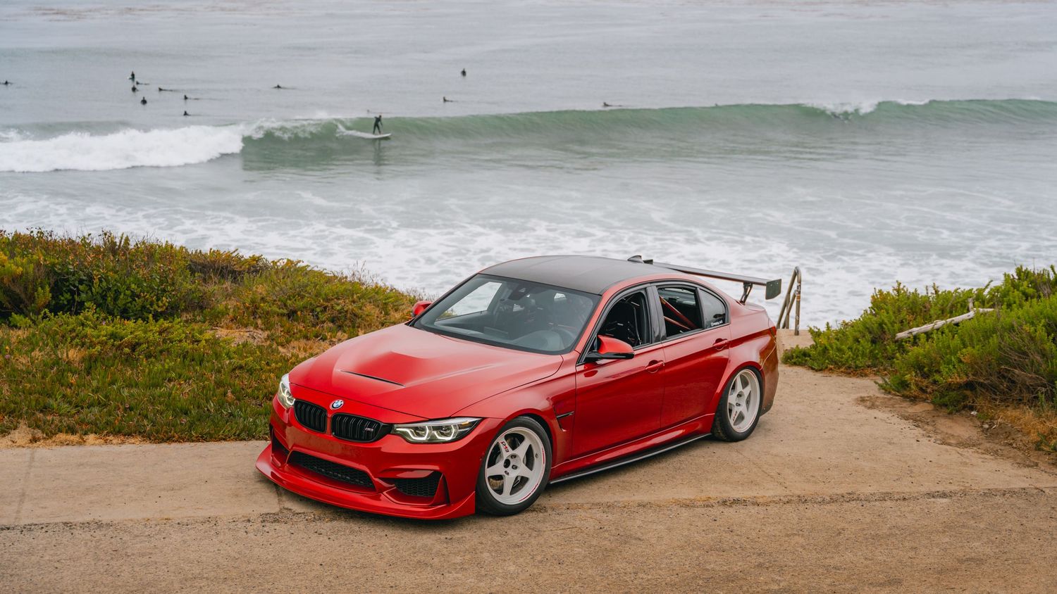 Red BMW M3 sedan parked near ocean overlook with surfers riding waves in the background on a cloudy day.