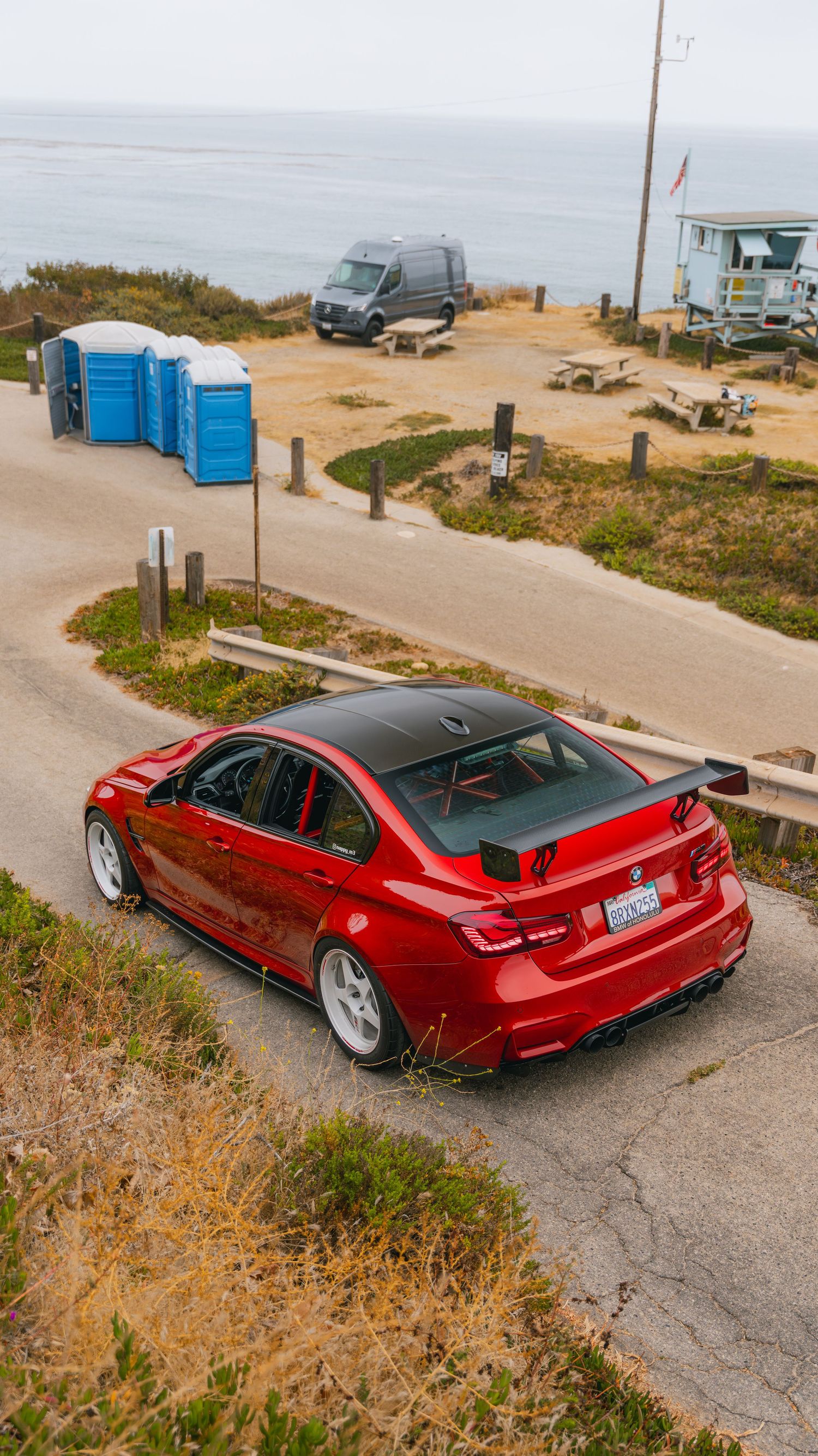 Red BMW M3 sedan with carbon fiber roof and spoiler parked near coastal overlook with portable toilets and van in background.