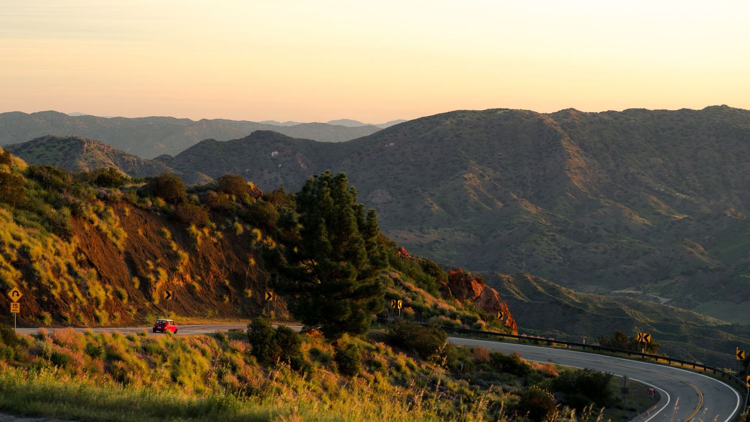 Winding mountain road curves through golden hills at sunset with mountains silhouetted against the warm sky.
