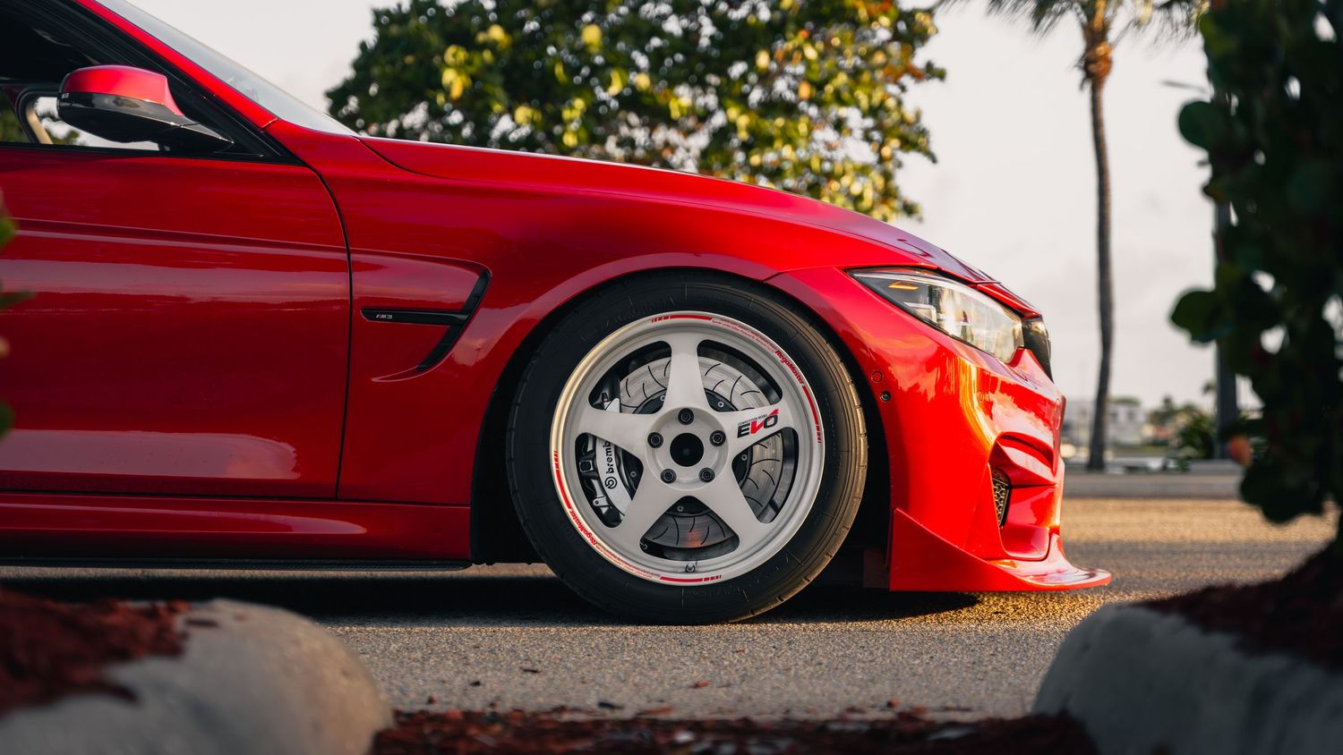Red BMW M4 with white aftermarket wheels on a sunny day, showcasing aggressive styling and low stance.