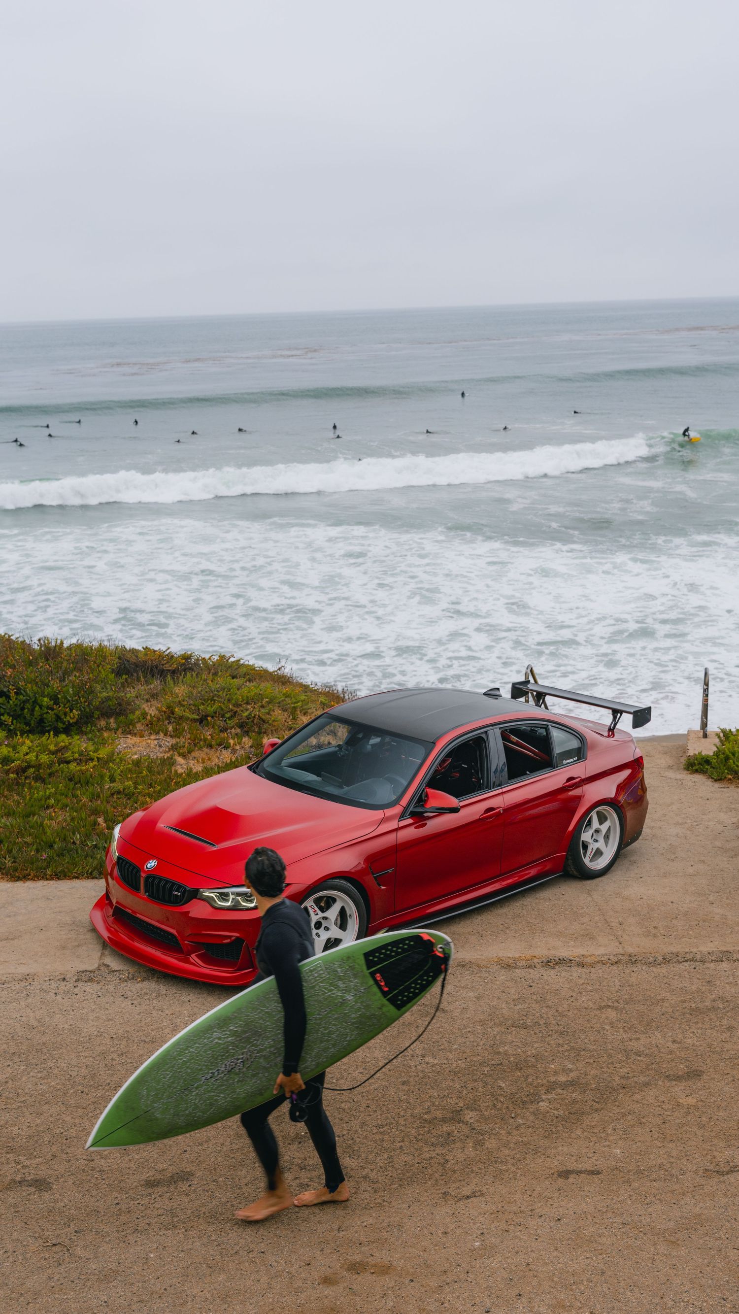 A surfer in a wetsuit carries a green surfboard past a modified red BMW M3 parked near a California beach.
