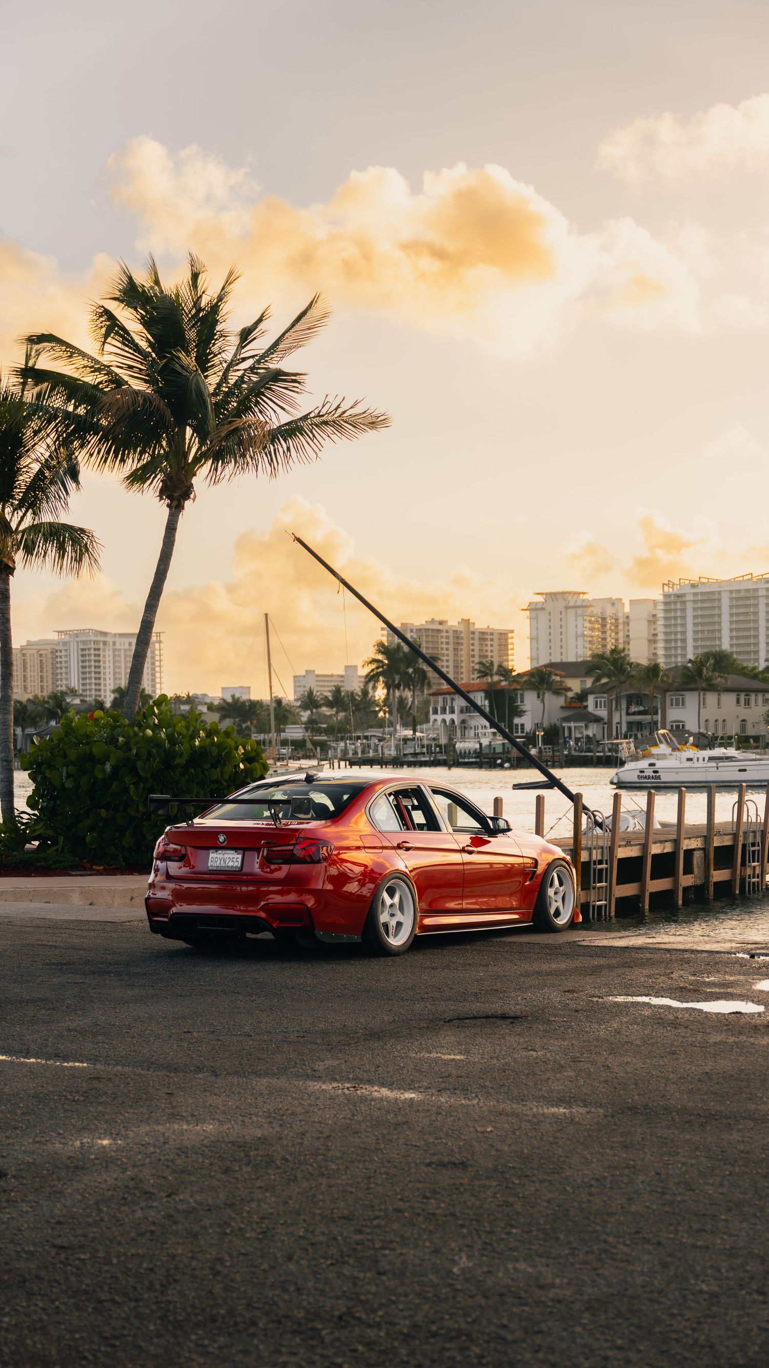 Red BMW sedan parked near a marina dock at sunset, with palm trees and city skyline in the background.