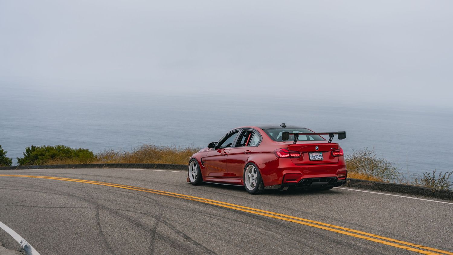Red BMW sedan with rear spoiler driving on a coastal highway with ocean views on a cloudy day.