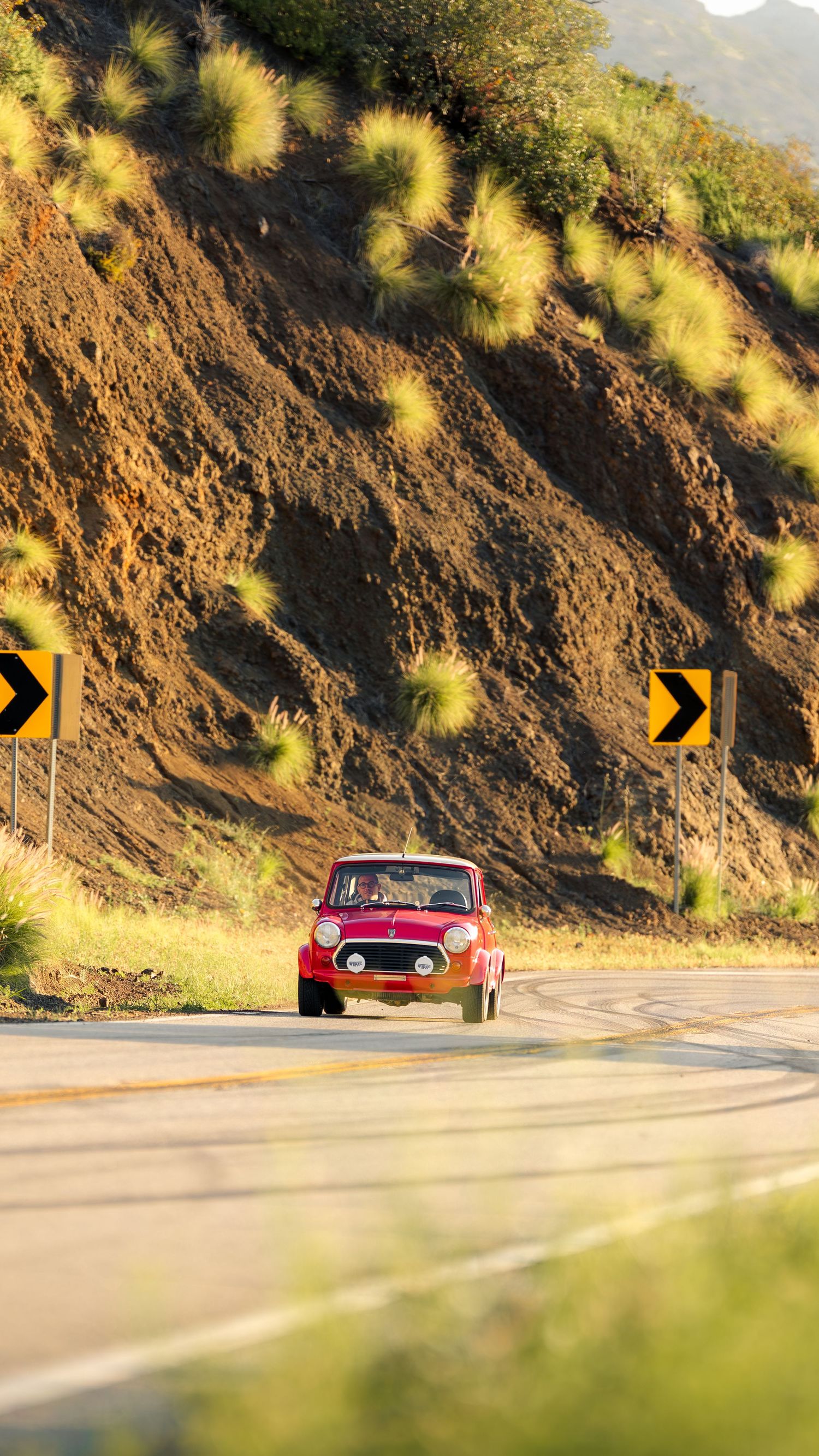 A vintage red car drives along a curving coastal road with warning signs and golden sunlit hillside.