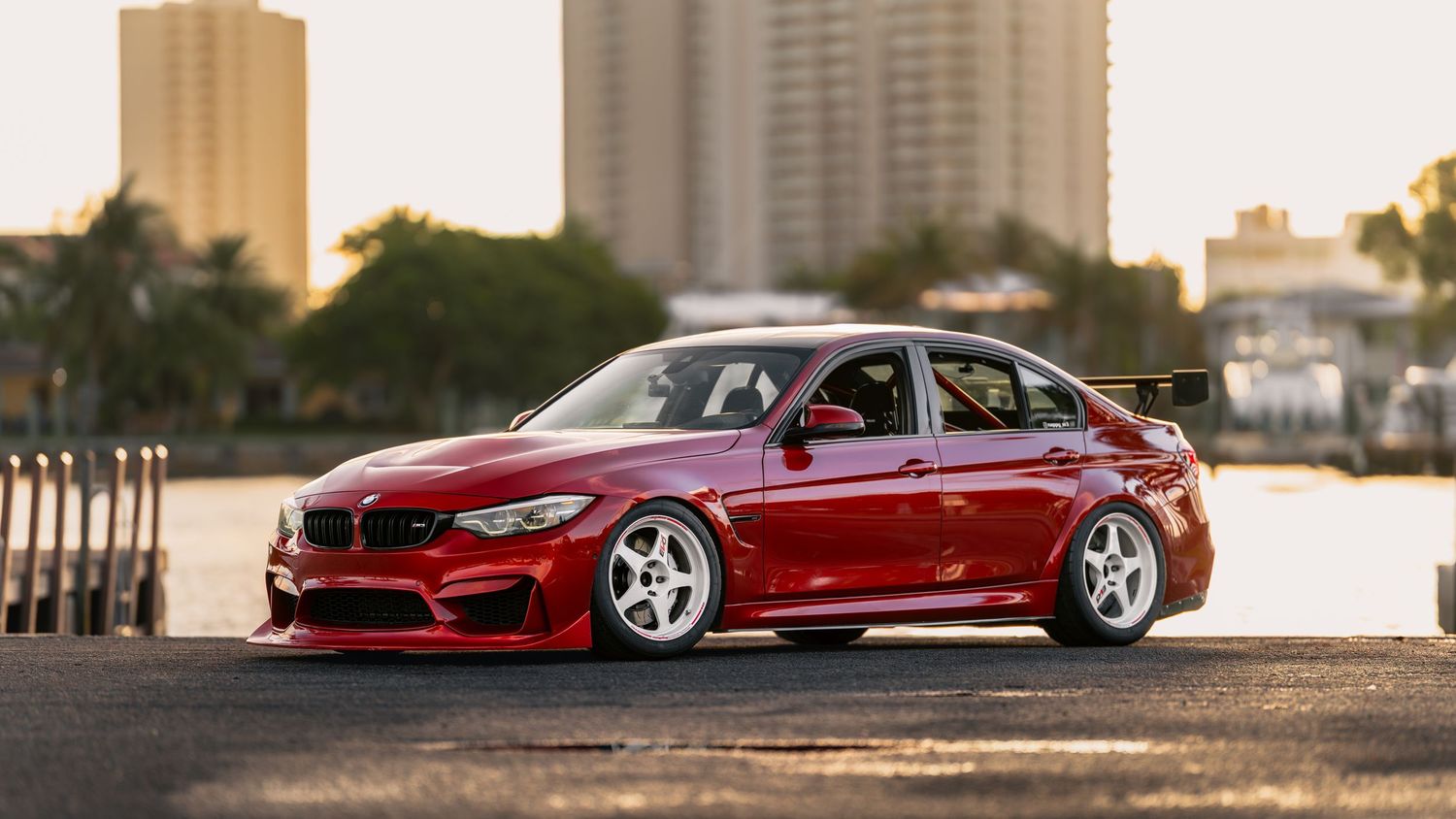 A red BMW M3 sports sedan with wide body kit and white racing wheels parked near waterfront buildings at sunset.