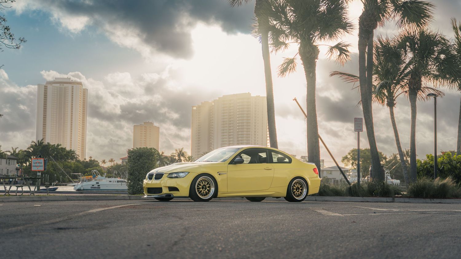 Yellow BMW M3 coupe parked on asphalt with palm trees and city skyline under dramatic cloudy sky.