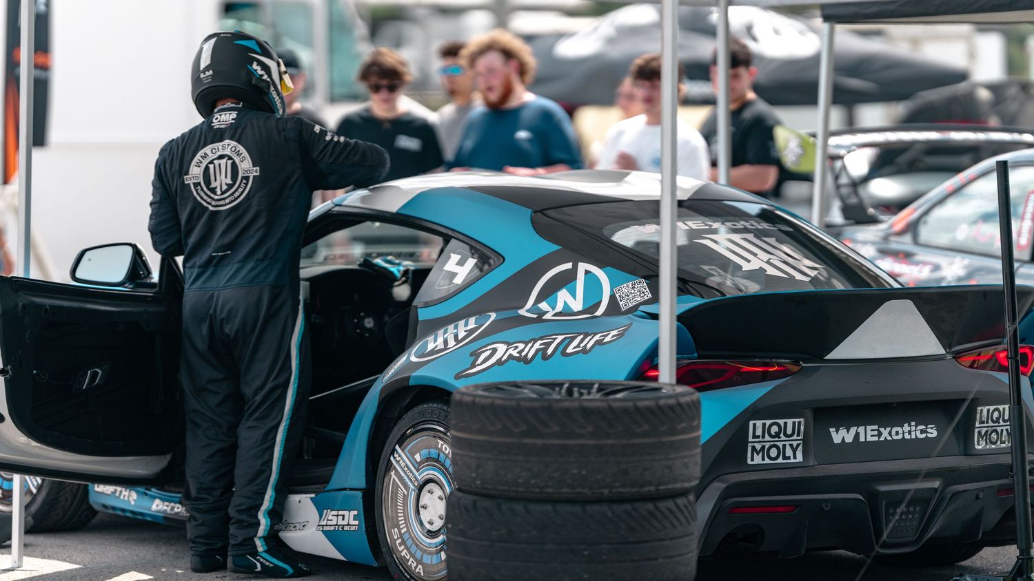 A racing driver in black overalls stands beside a teal drift car with sponsor logos at a motorsport event.