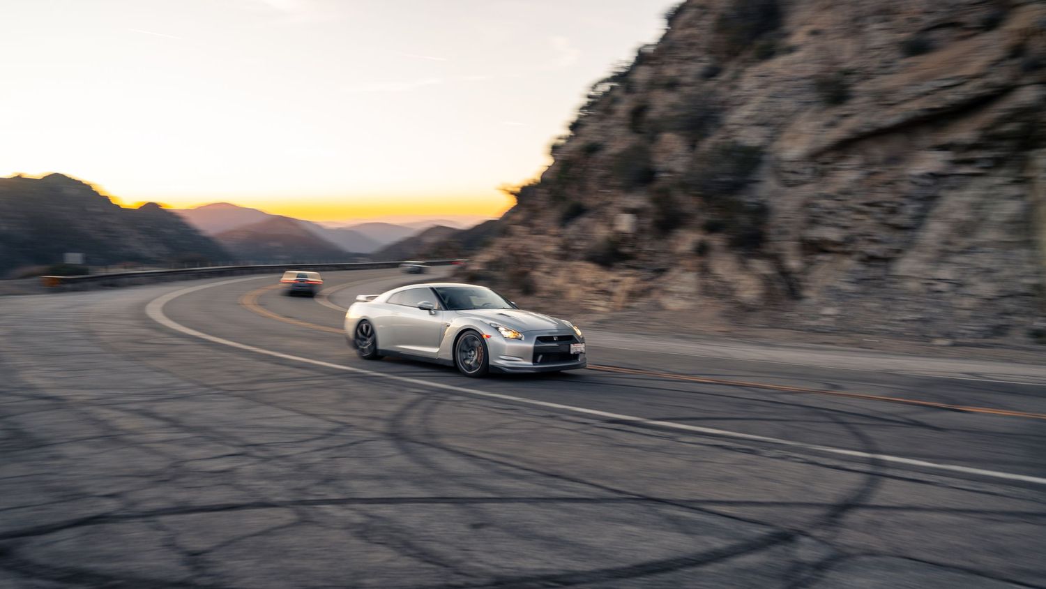 Black tire marks streak across a winding mountain road with sports cars in view.