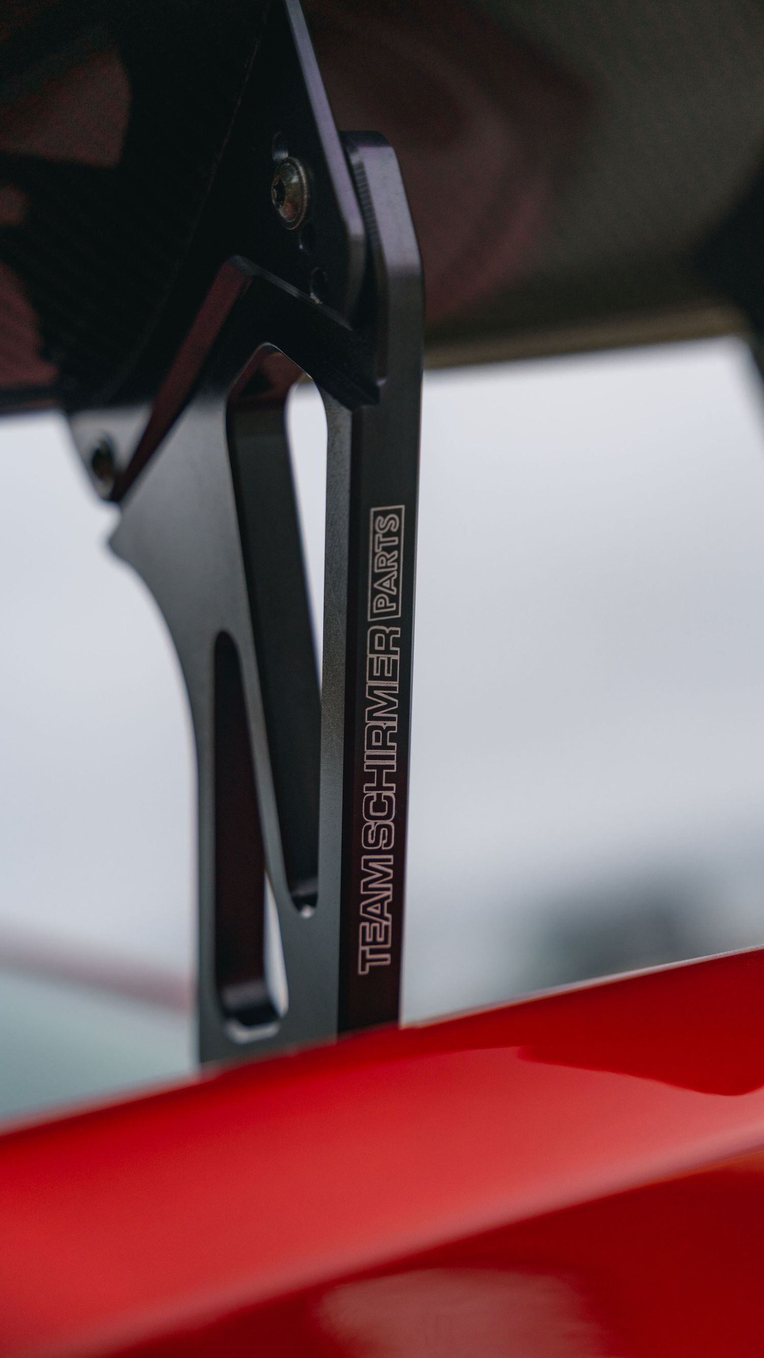Close-up of a black Team Schirmer Parts bracket mounted on a red car body, showcasing precision engineering.