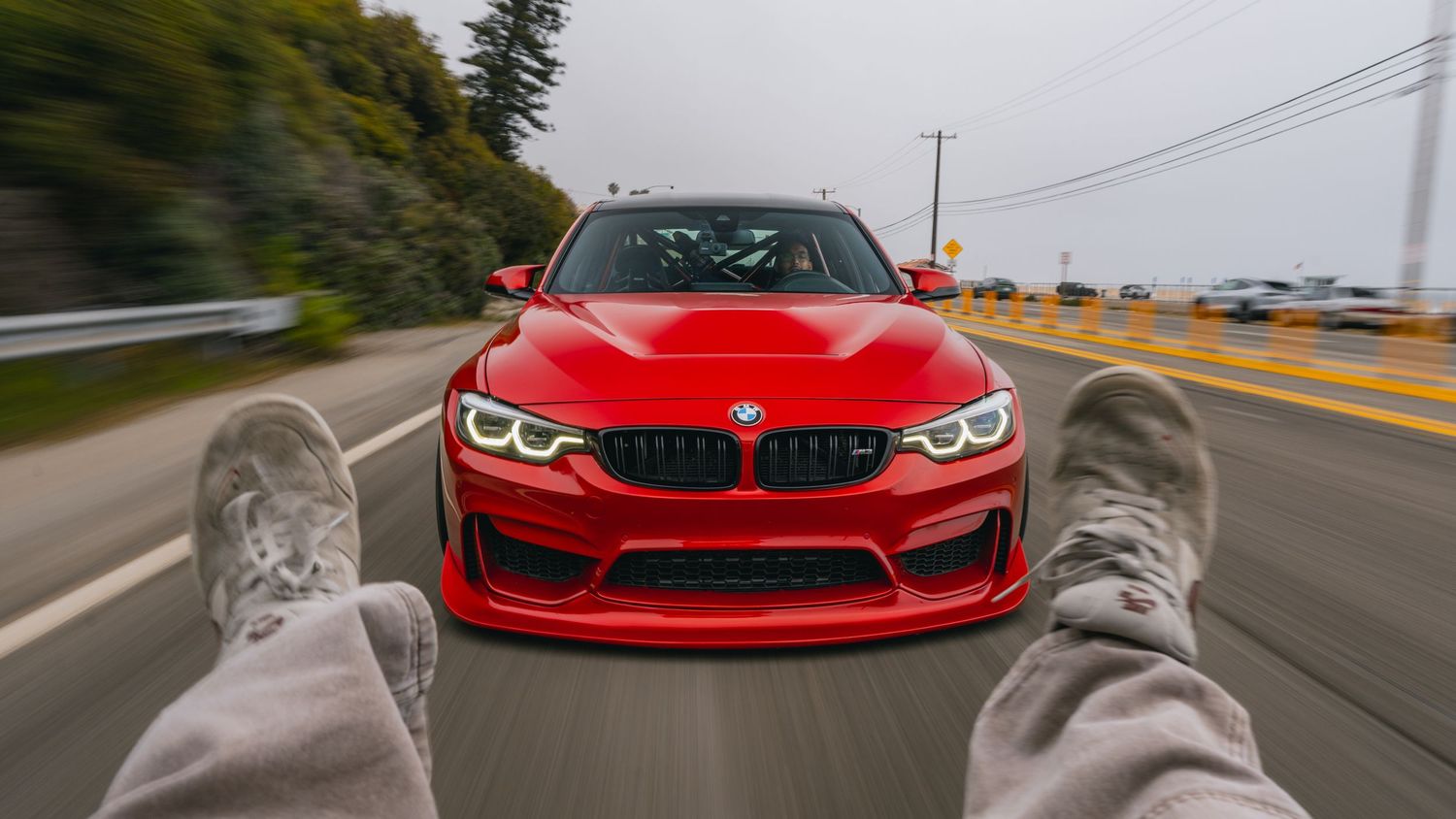Red BMW M3 speeding on highway shot from hood perspective with feet visible in foreground.