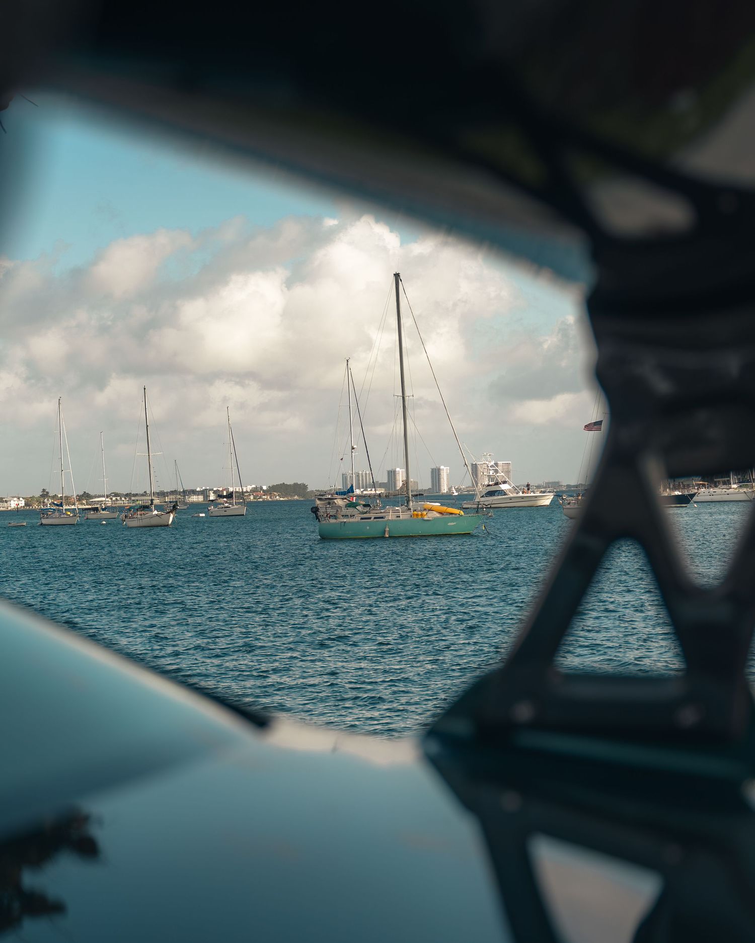 Sailboats moored in a calm harbor viewed through a boat window with city skyline and cloudy sky in background.