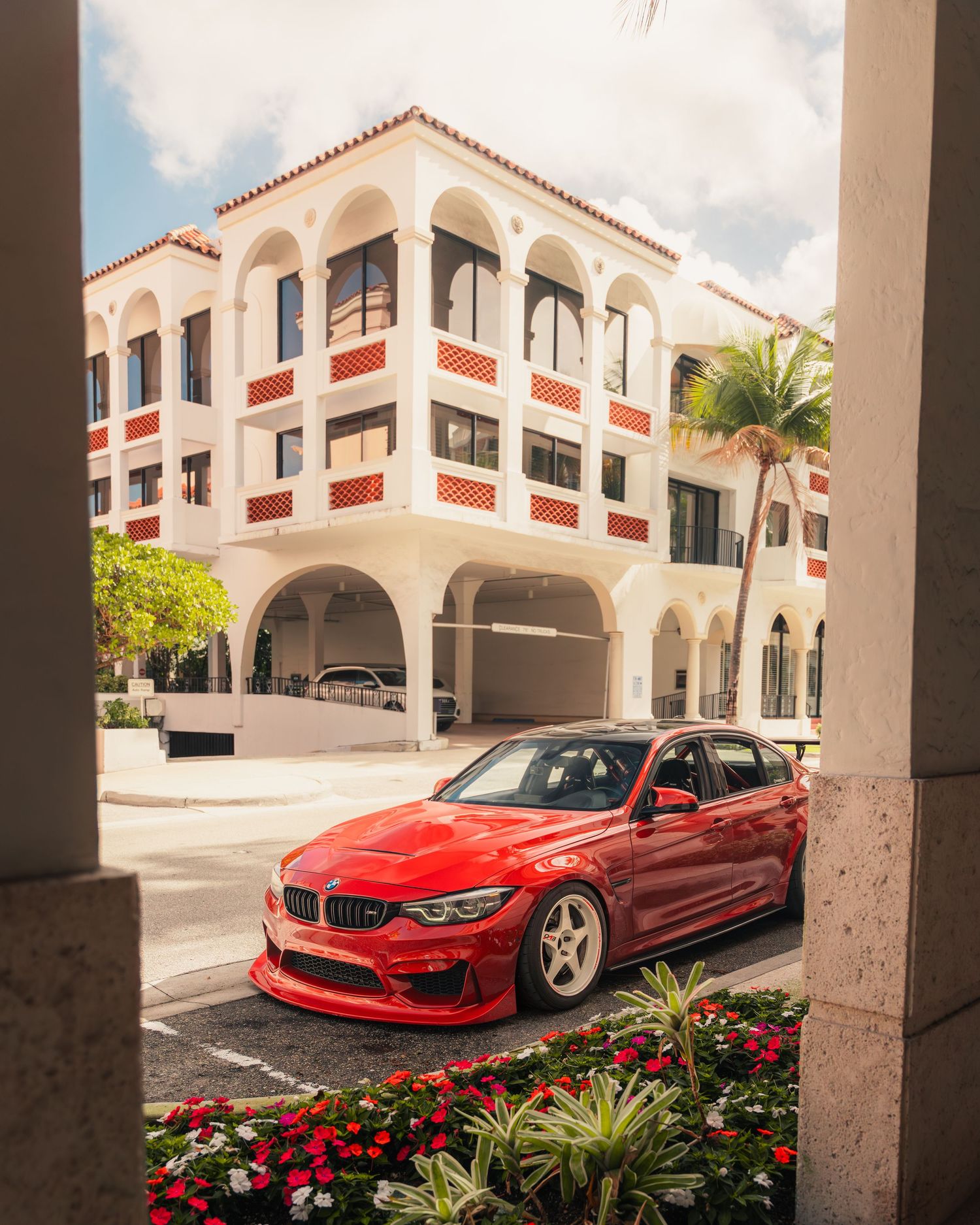 A red BMW M3 with custom wheels parked in front of a white Spanish-style building with red accents and palm trees.