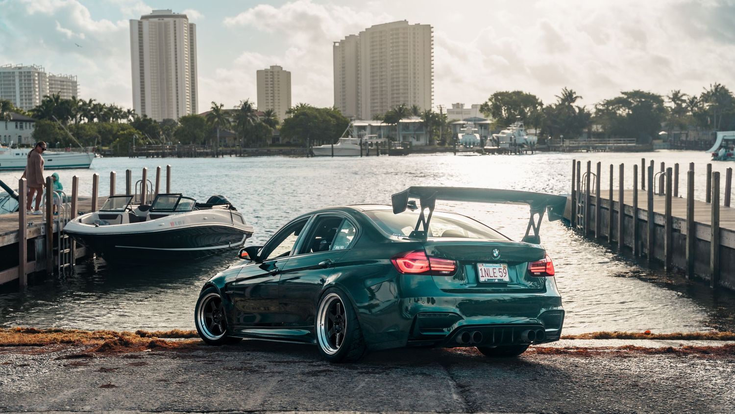 A dark green modified BMW M3 with large rear spoiler parked on a waterfront dock with boats and city skyline.