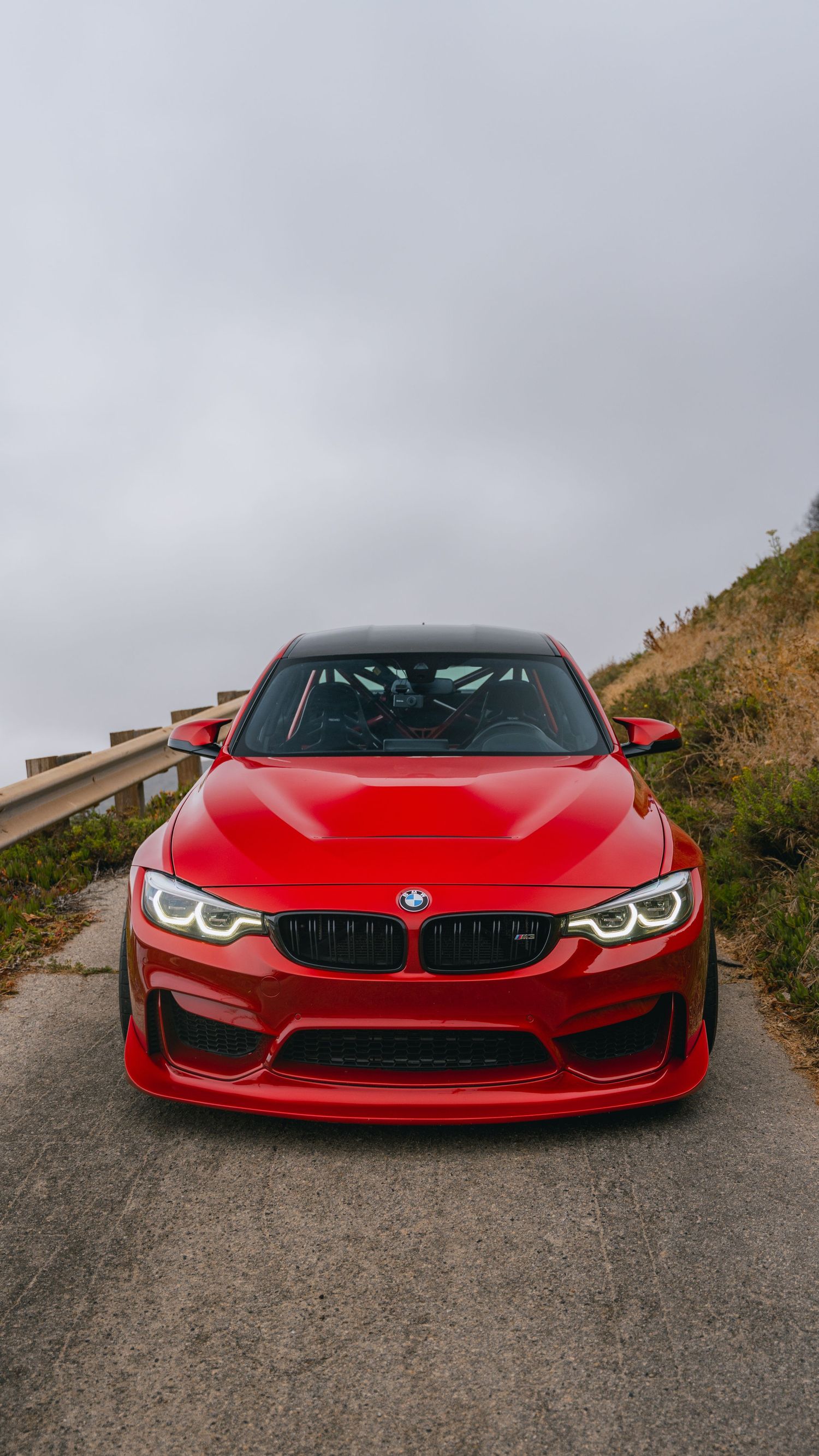 Front view of a red BMW M3 with aggressive body kit parked on a hillside road under cloudy skies.