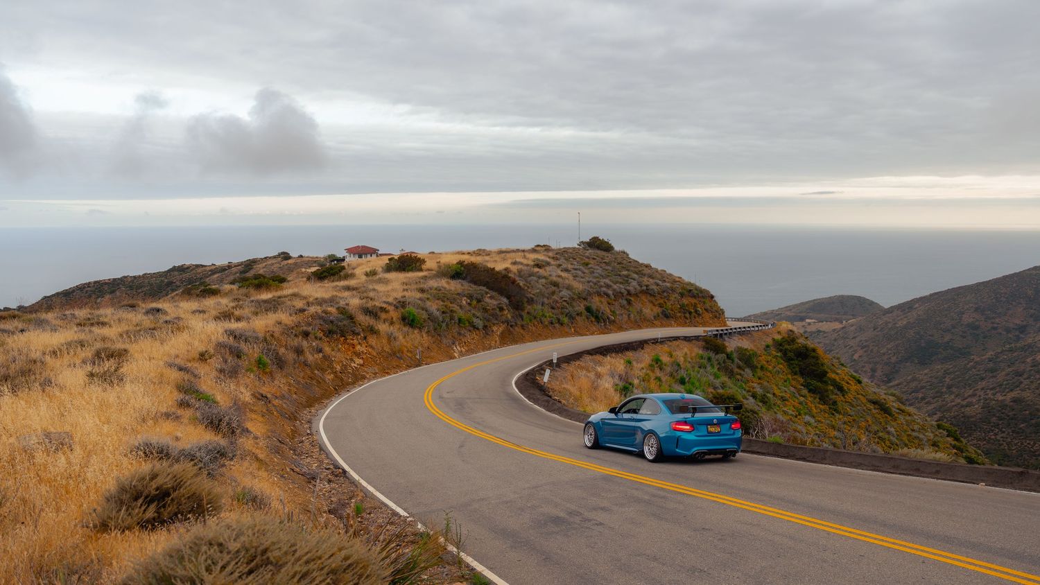 A blue BMW sedan drives along a winding coastal mountain road under an overcast sky with ocean views.
