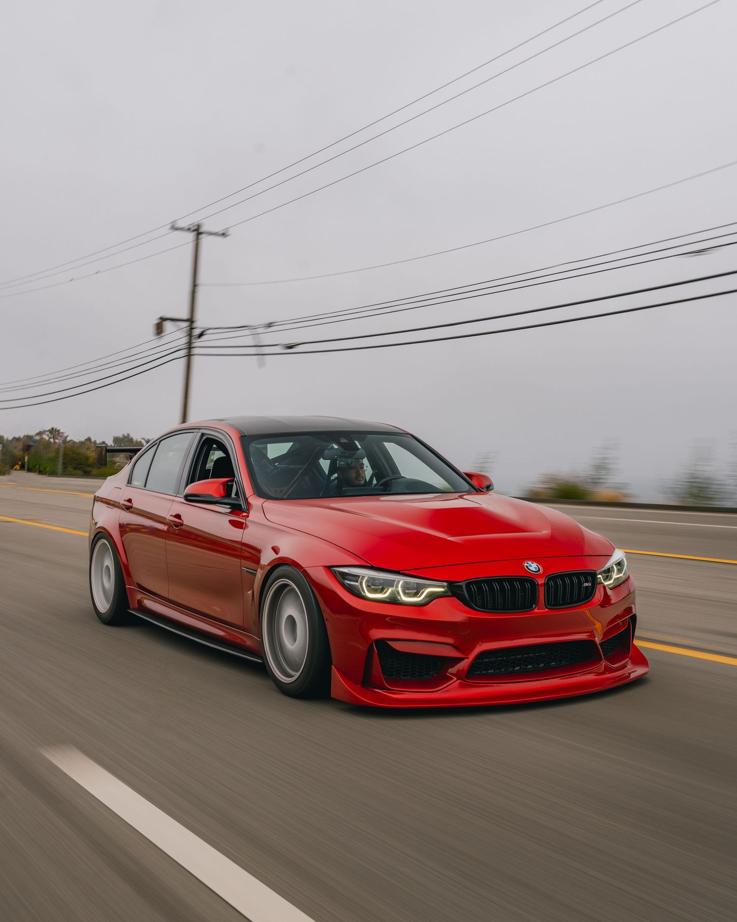 Modified red BMW M3 sedan with wide body kit driving on coastal highway.