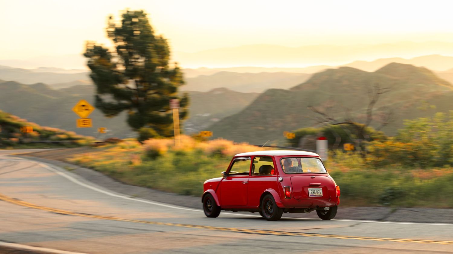 A classic red sports car drives along a winding mountain road during golden hour sunset.