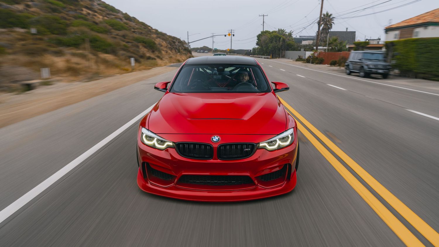 Red BMW M4 driving on a scenic highway road, front view showing aggressive styling and iconic kidney grille design.