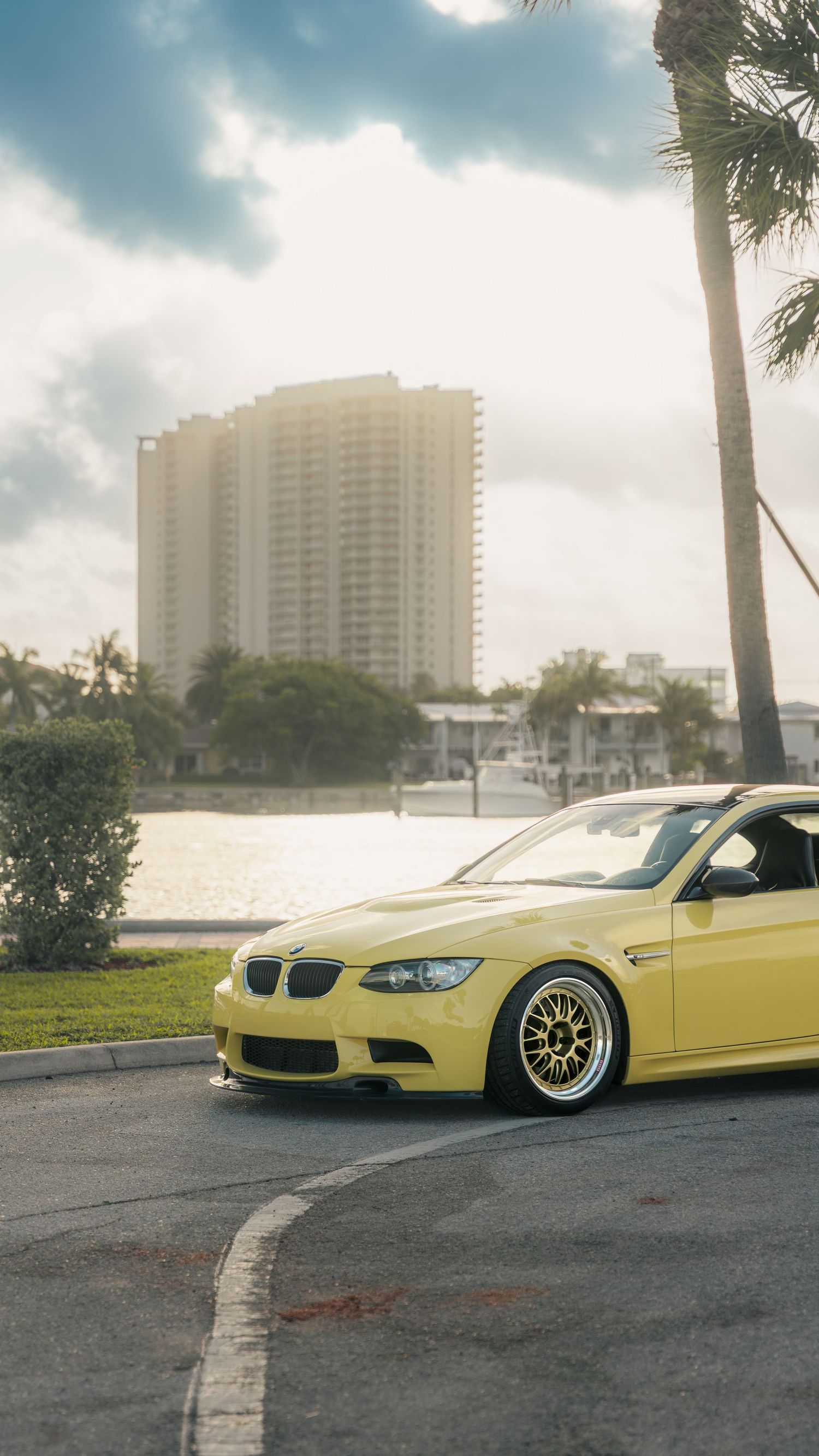 Yellow BMW M3 E92 slammed on BBS wheels parked near waterfront with palm trees and high-rise building in background.
