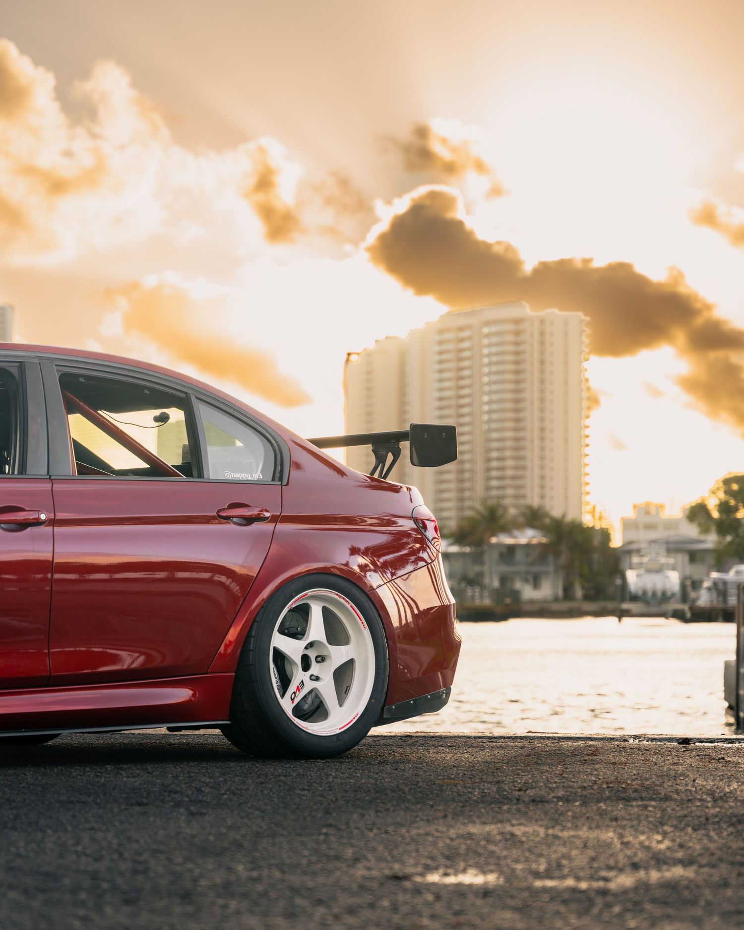 Red modified Mitsubishi Lancer Evo with white wheels and rear spoiler parked at sunset with city skyline backdrop.
