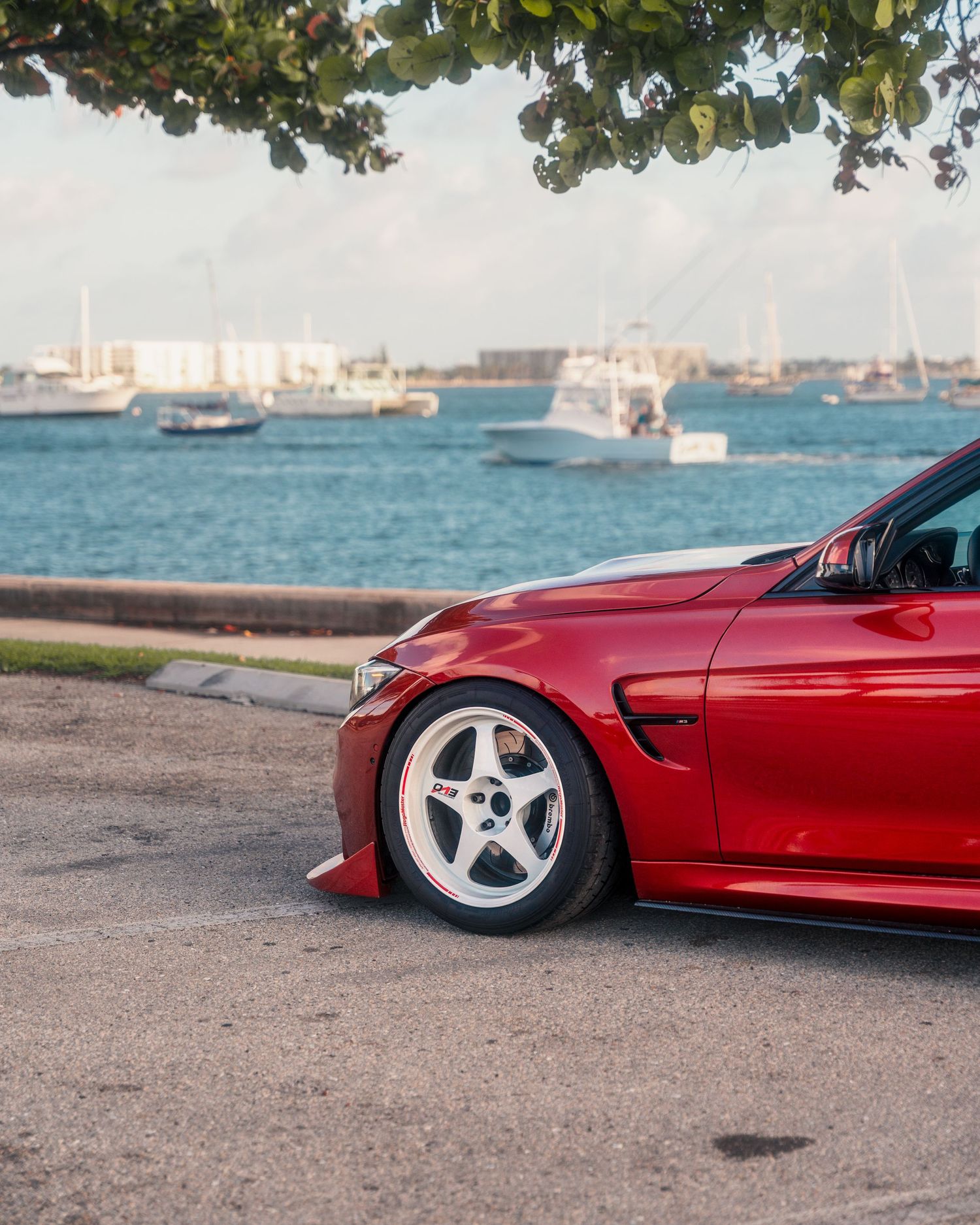 Red BMW M4 on white aftermarket wheels parked near waterfront with boats and blue water in background.