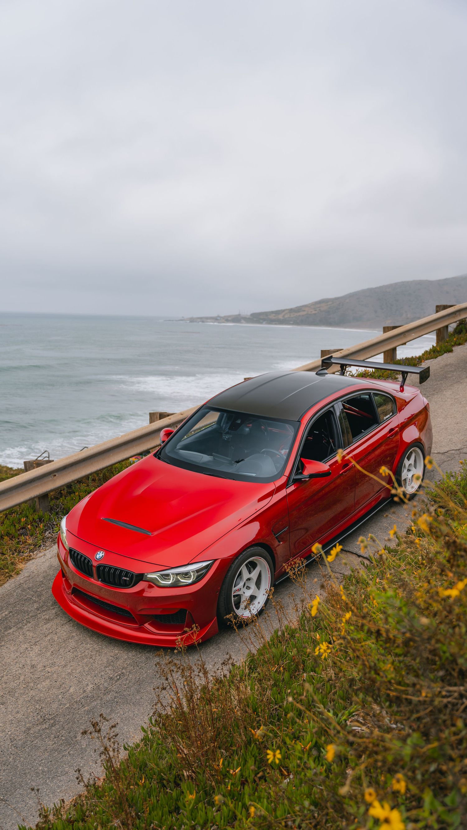 Red BMW M4 with carbon roof and rear wing parked on coastal highway overlooking ocean on overcast day.