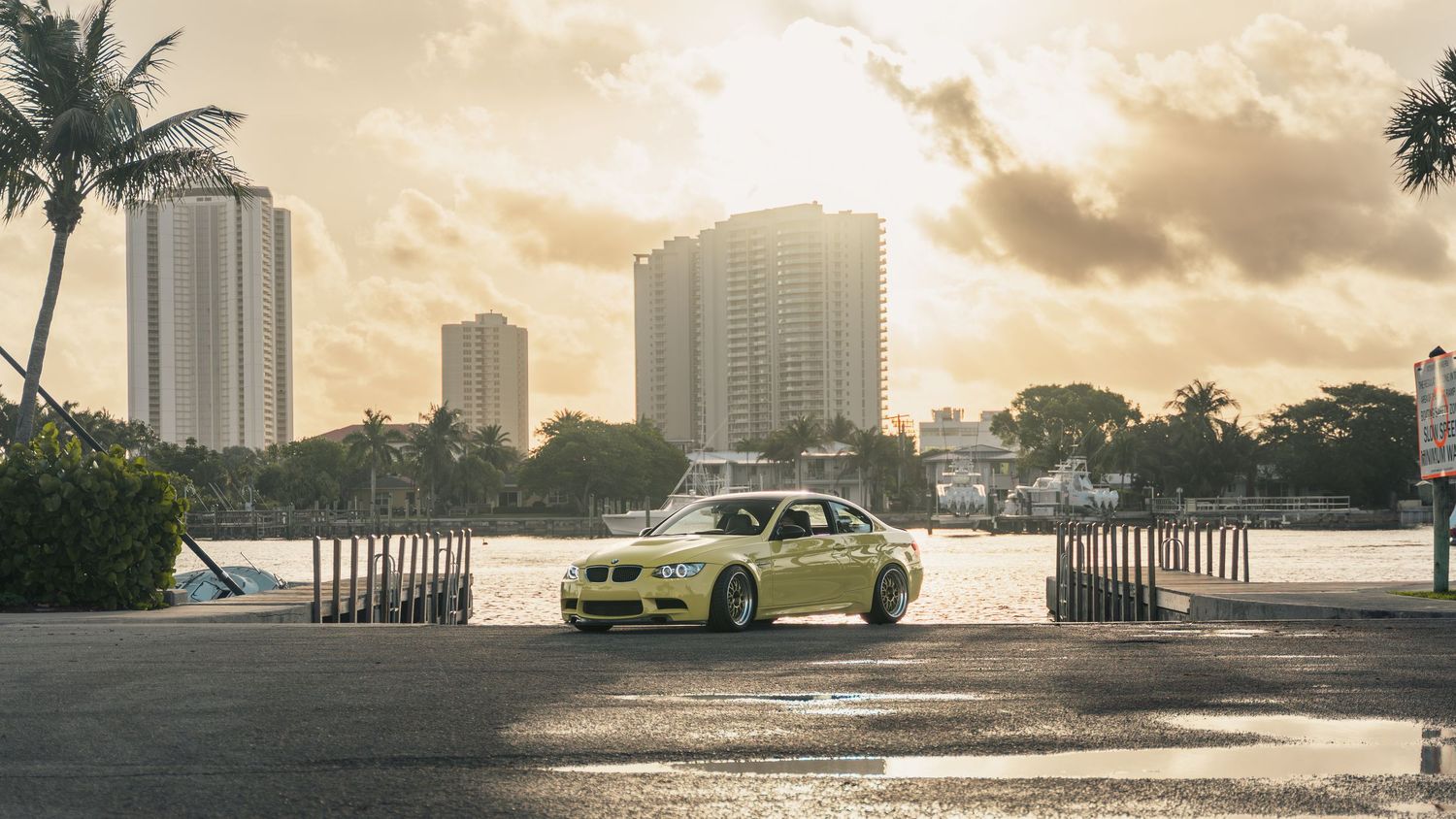 Yellow BMW M3 parked near waterfront with palm trees and city skyline under dramatic cloudy sky.