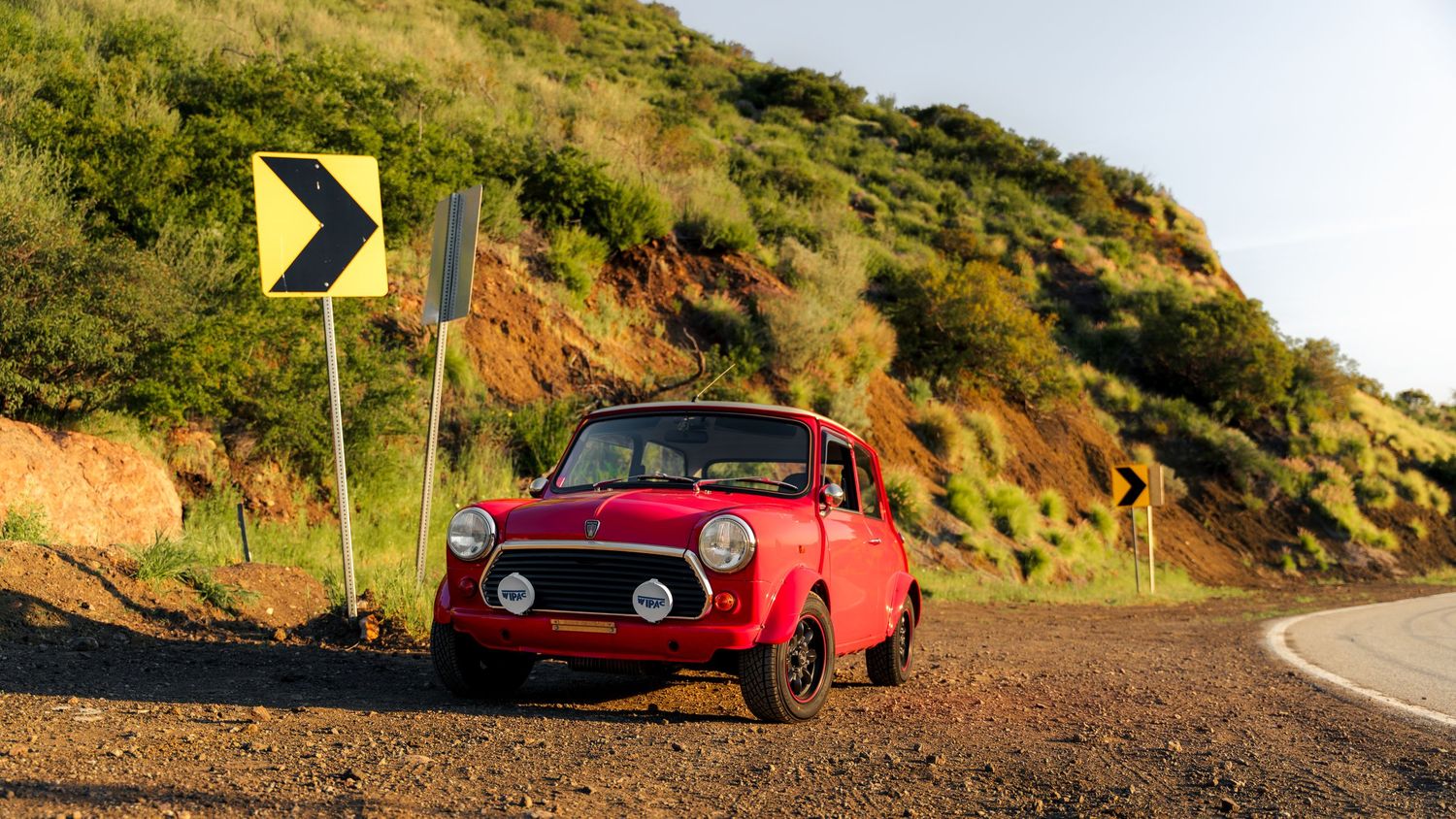 A red vintage car drives along a winding coastal mountain road with yellow signs visible in the distance.