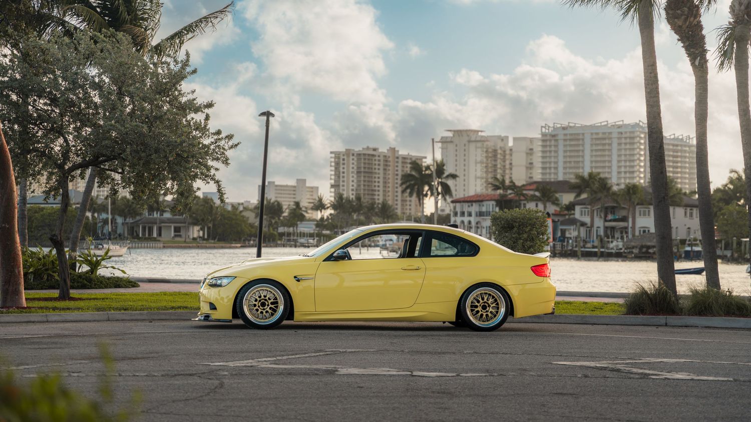 Yellow BMW M3 coupe on lowered suspension with gold wheels parked near waterfront palm trees and city buildings.