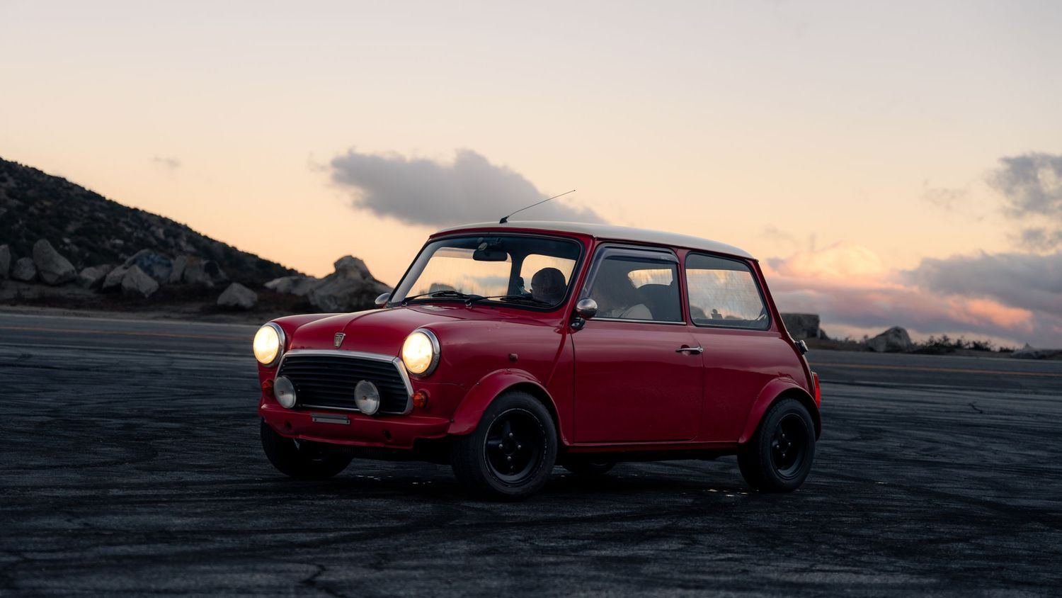 Classic red Mini Cooper with black accents parked against sunset sky.