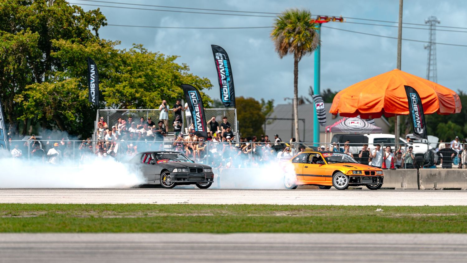 Two cars drifting with massive smoke clouds at an outdoor motorsport event with spectators and palm trees.