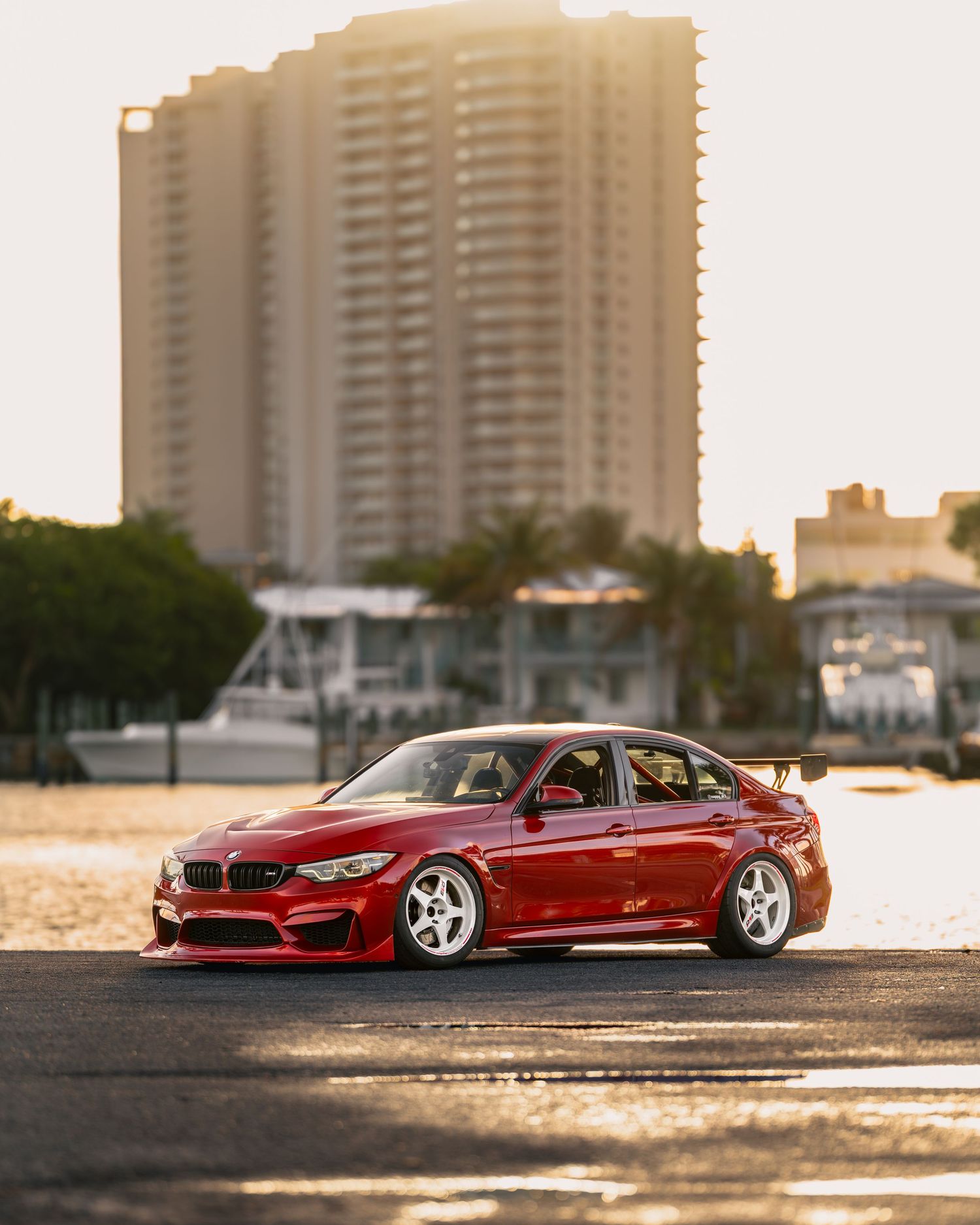 Red BMW M3 sedan with wide body kit and white wheels parked near waterfront with luxury high-rise building at sunset.