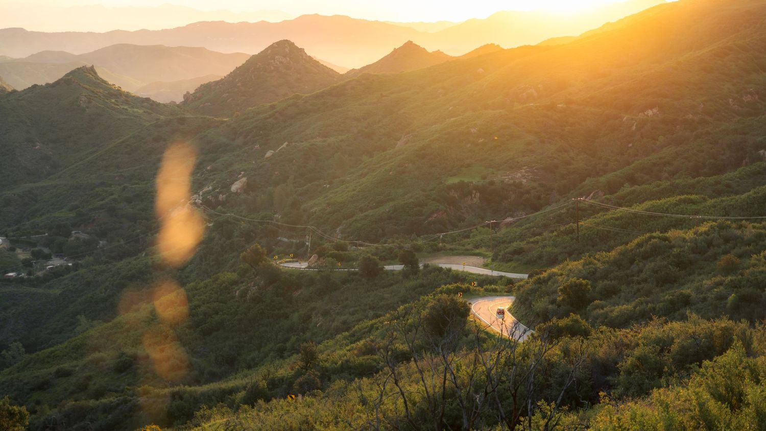 Winding mountain road curves through green hills at golden sunset with sunbeams in California's coastal range.