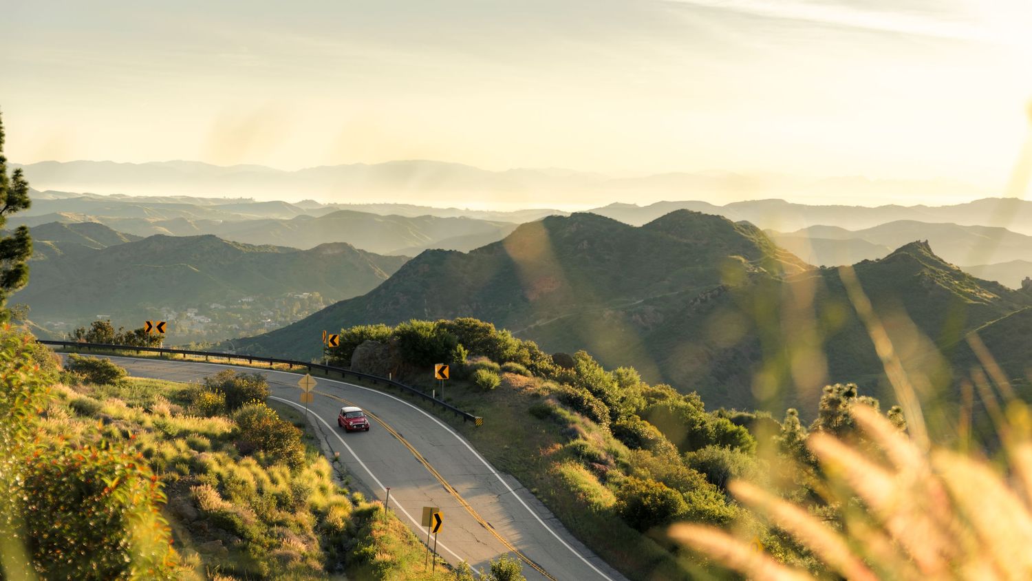 Scenic winding mountain road curves through golden hills with distant mountain ranges at sunset.