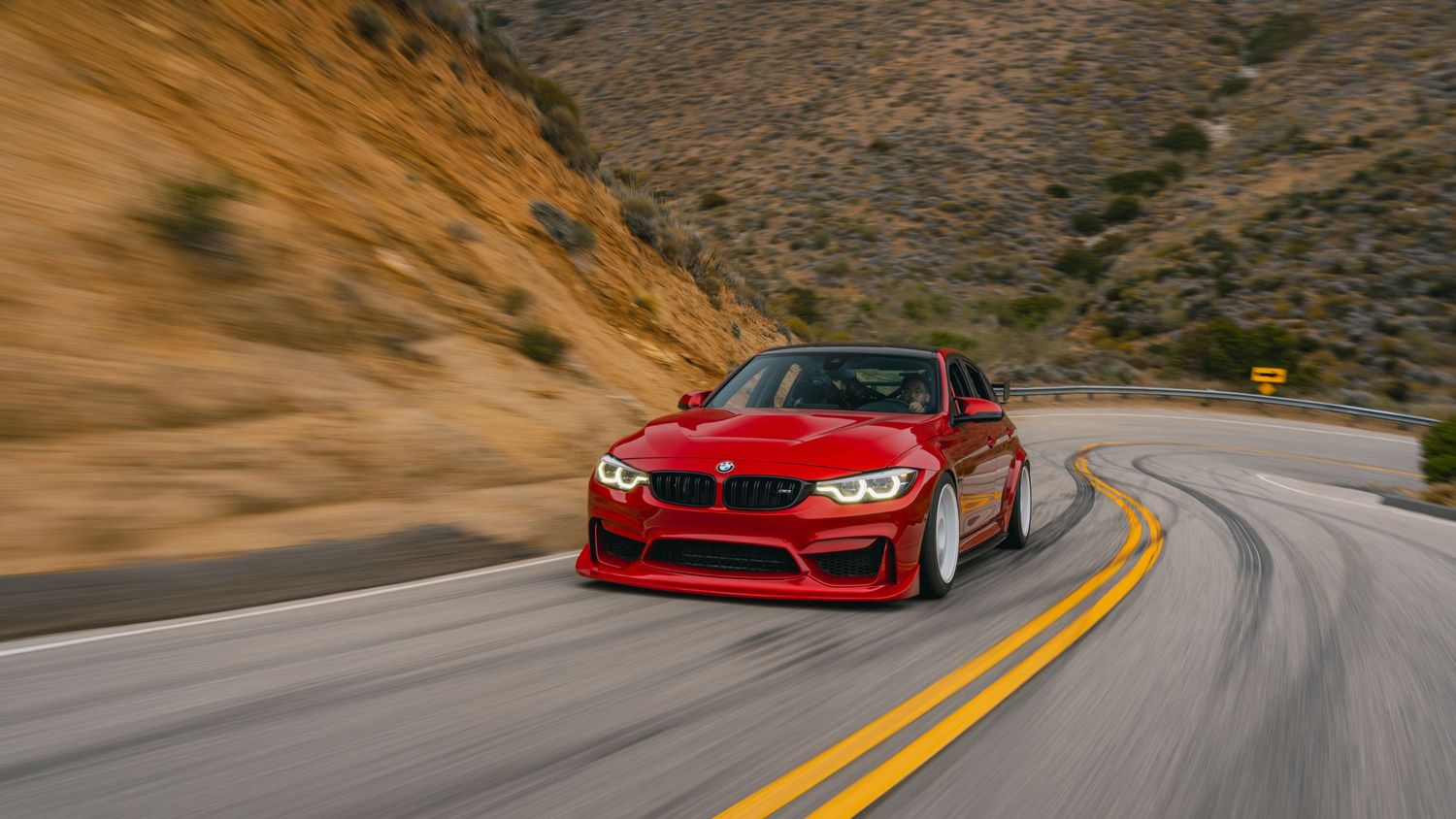 Red BMW M3 speeding on a winding mountain road with rocky hills in the background.