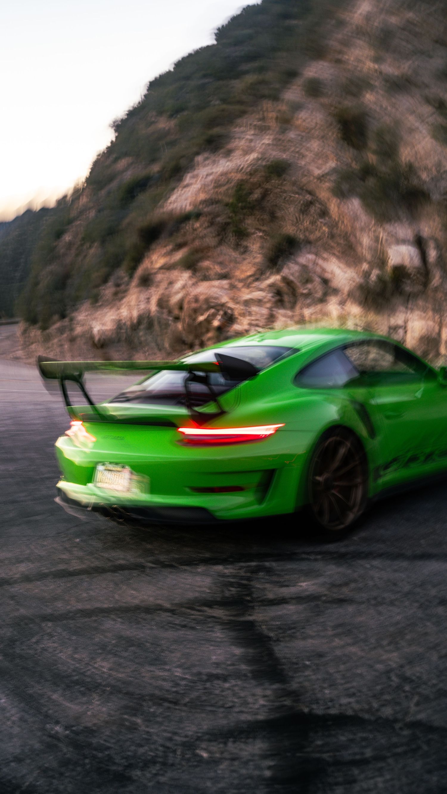 Neon green Porsche 911 GT3 RS with large rear wing driving on mountain road at dusk.