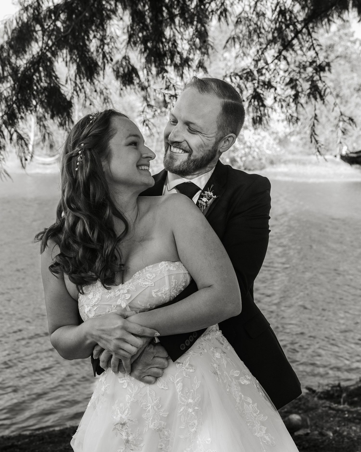 Black and white photo of a smiling bride and groom embracing near a waterfront under hanging tree branches.