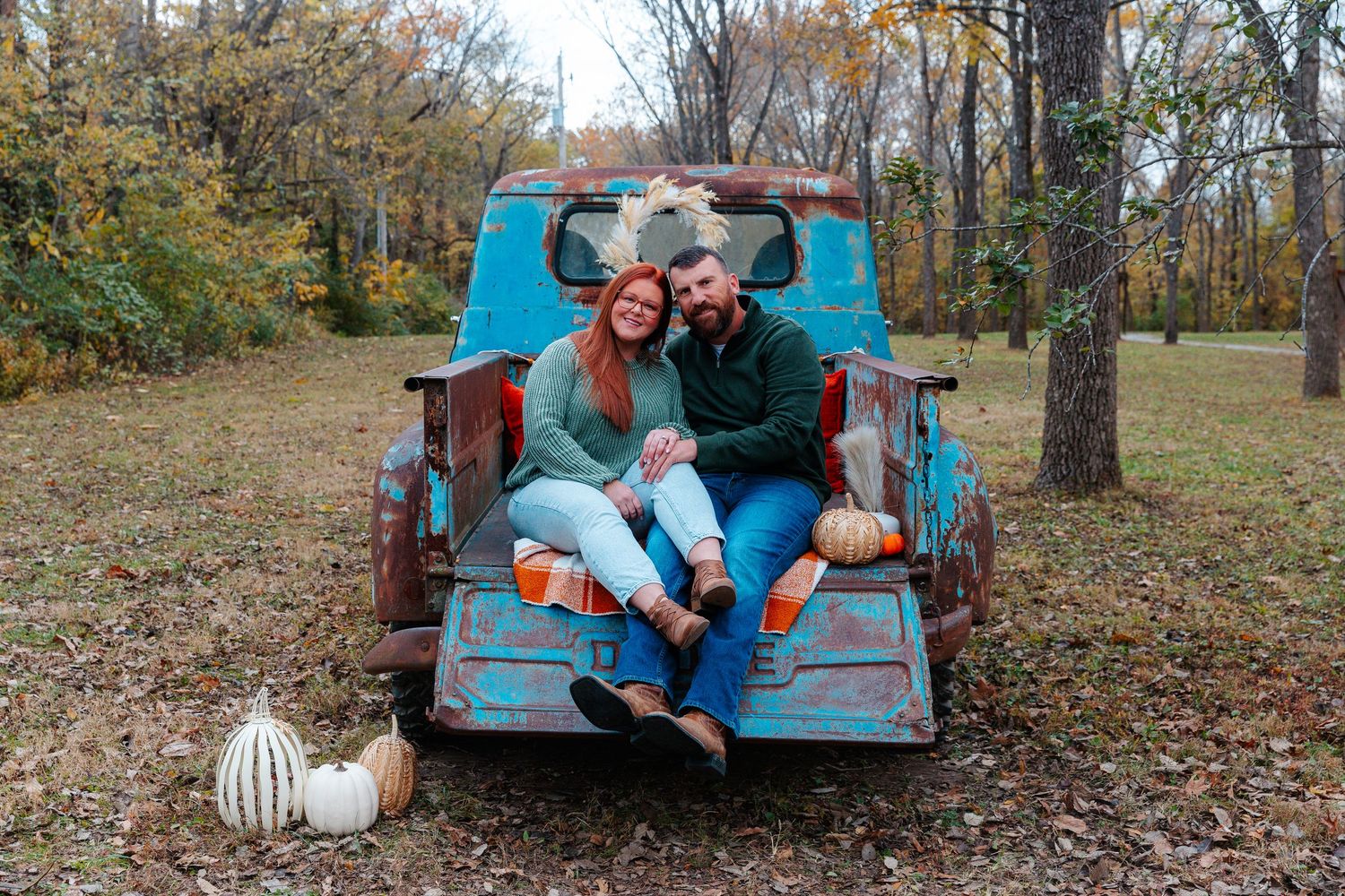 Couple sitting in rustic blue truck bed with pumpkins during fall foliage season.