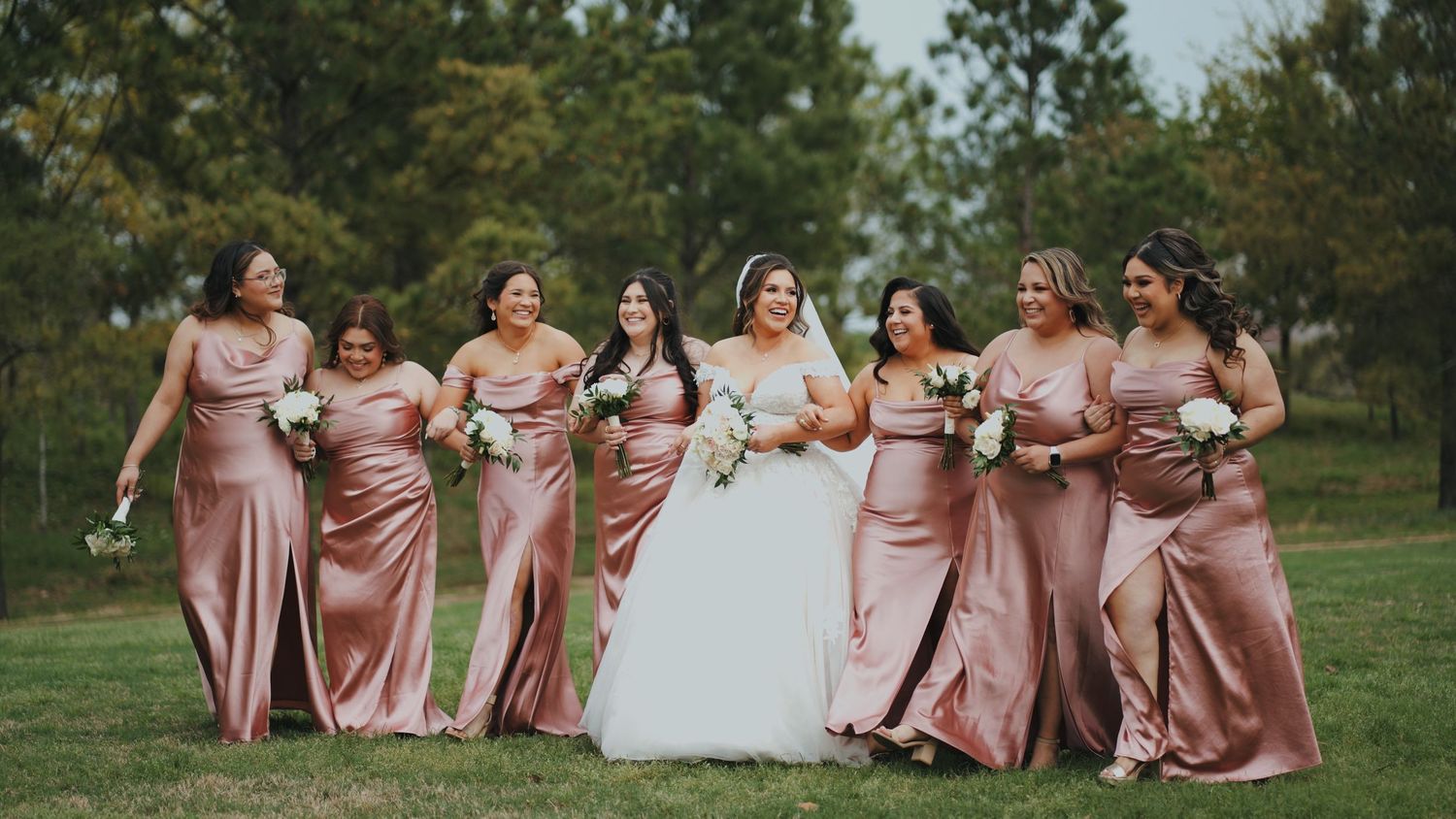 Wedding party in dusty rose dresses poses together outdoors on a sunny day in a garden setting.