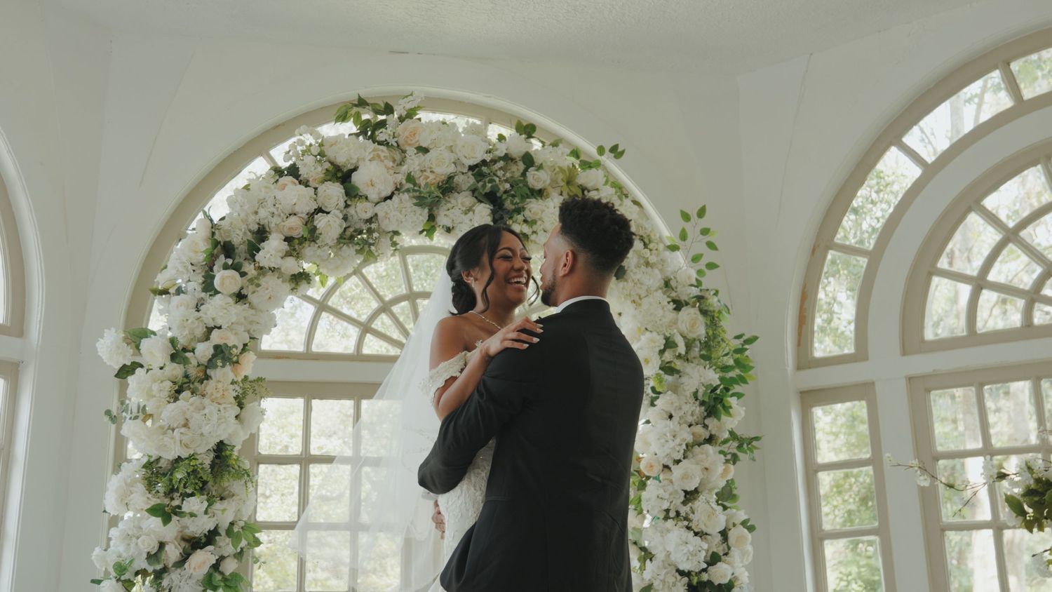 White floral arch decorations adorn a wedding ceremony space with arched windows.