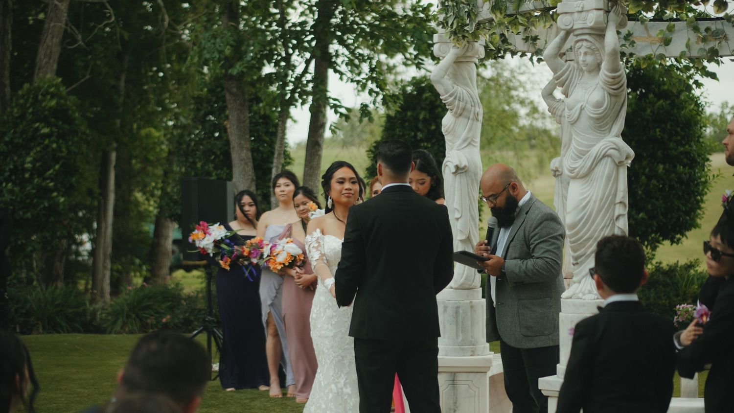 Outdoor wedding ceremony in progress among trees with white decorative pillars.