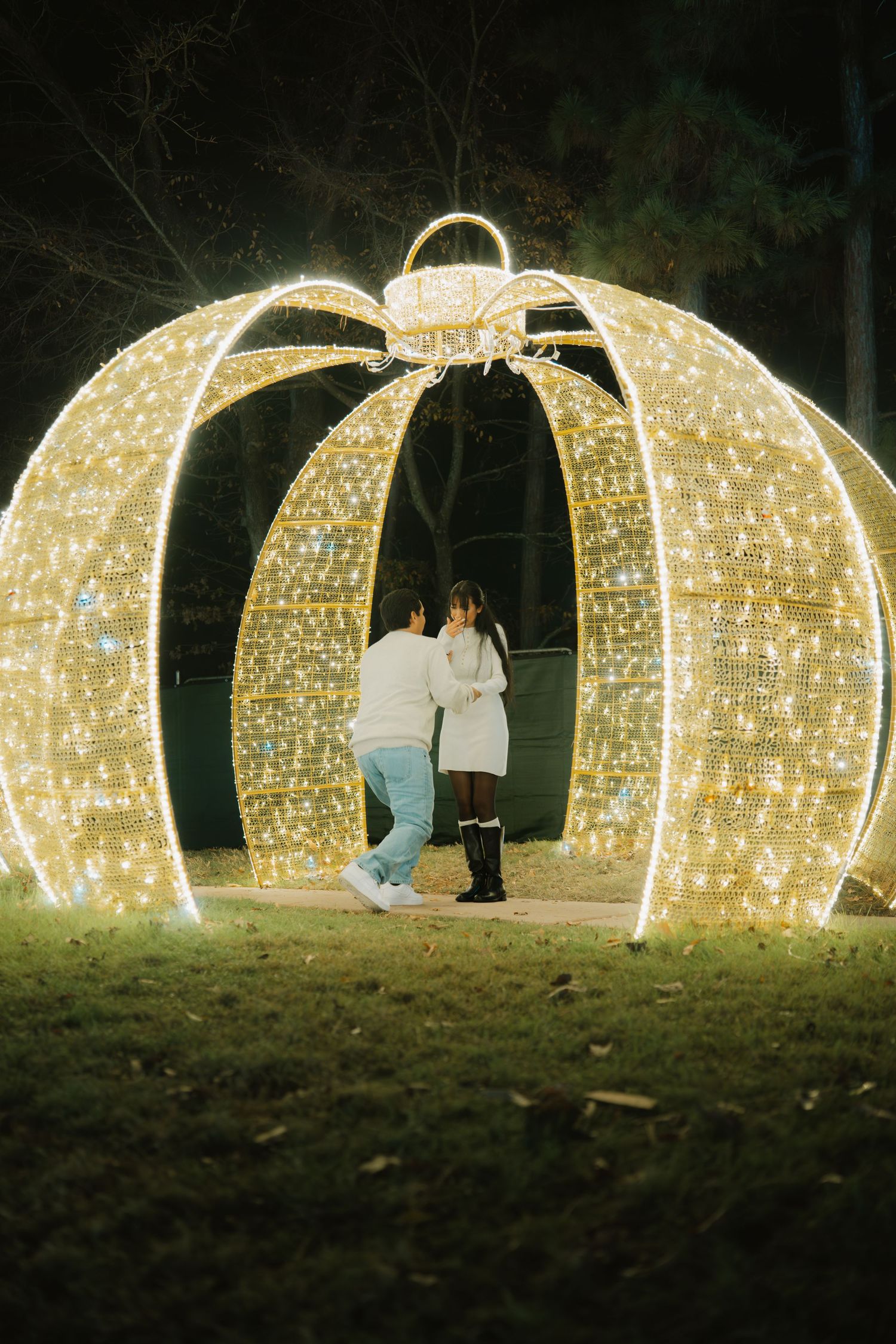 A couple shares a romantic moment inside a glowing pumpkin-shaped light installation in a dark outdoor setting.