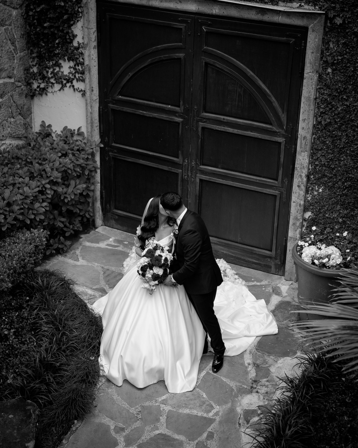 Black and white photo of a couple in wedding attire embracing by an arched wooden door.