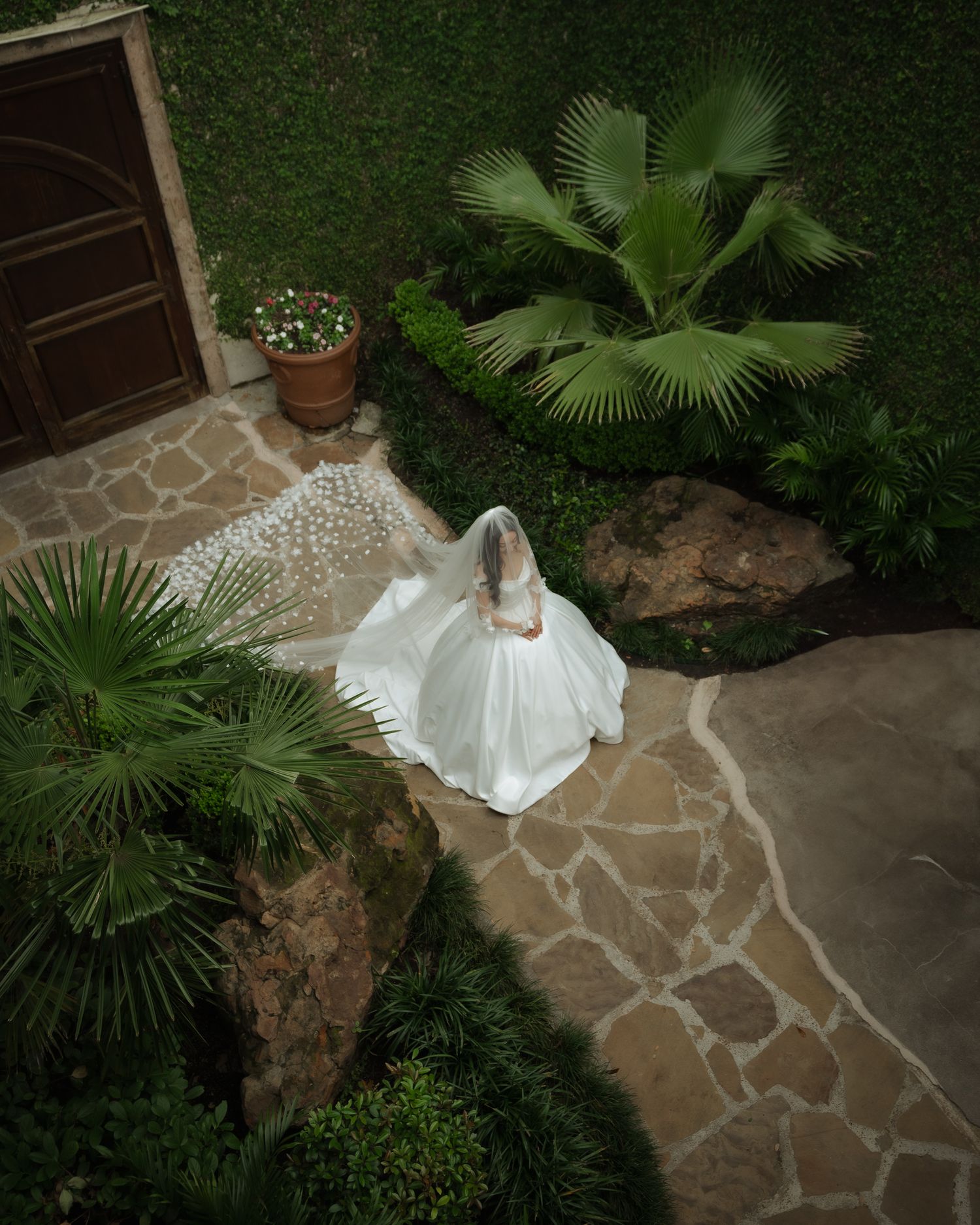 Overhead view of a flowing white wedding dress on a stone pathway surrounded by palm trees and garden landscaping.