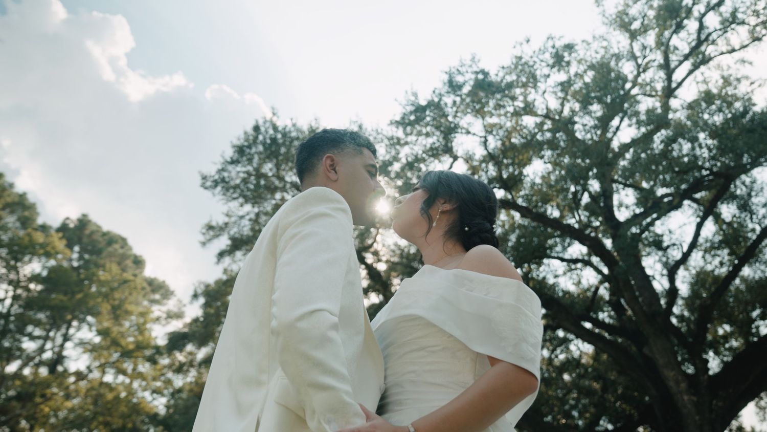 A couple in white wedding attire walks hand in hand through a lush garden setting during a romantic outdoor ceremony.