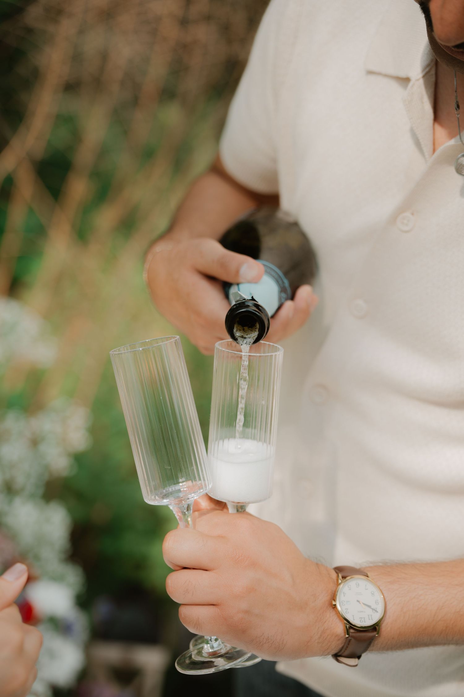 Hands pouring champagne into elegant glass flutes outdoors.