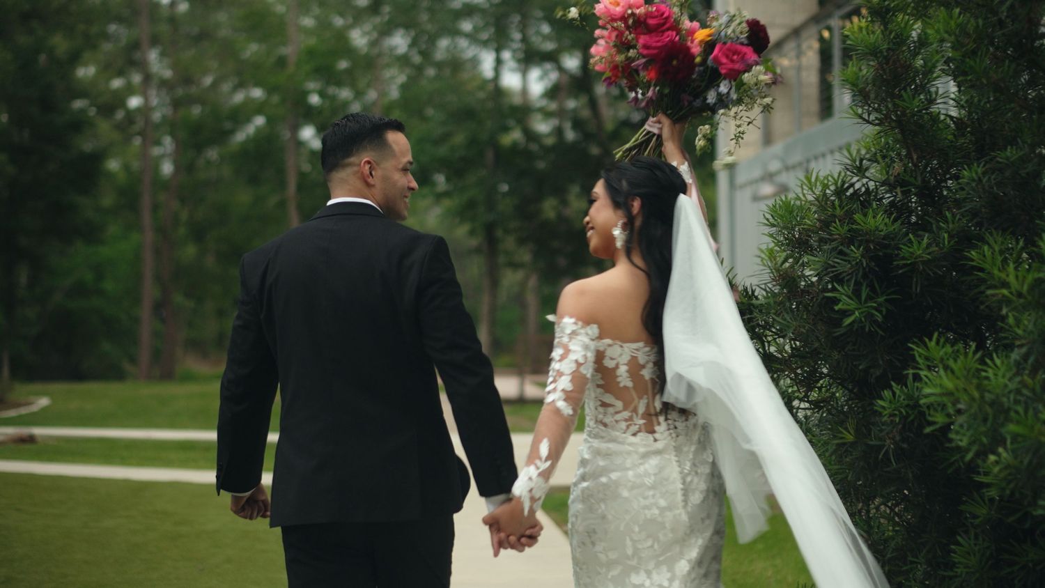 Wedding couple poses outdoors with vibrant red floral bouquet against lush green backdrop.