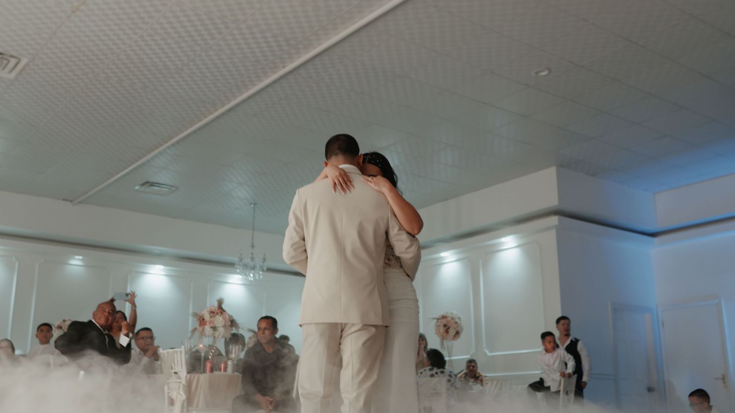 White wedding reception hall with elegant ceiling decor and hanging lights.