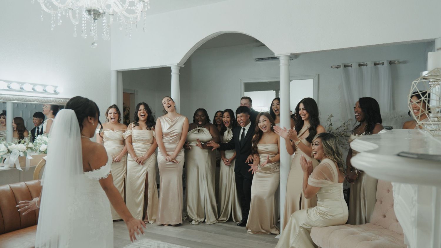Wedding party members in white dresses and black suits gather in an elegant bridal suite.